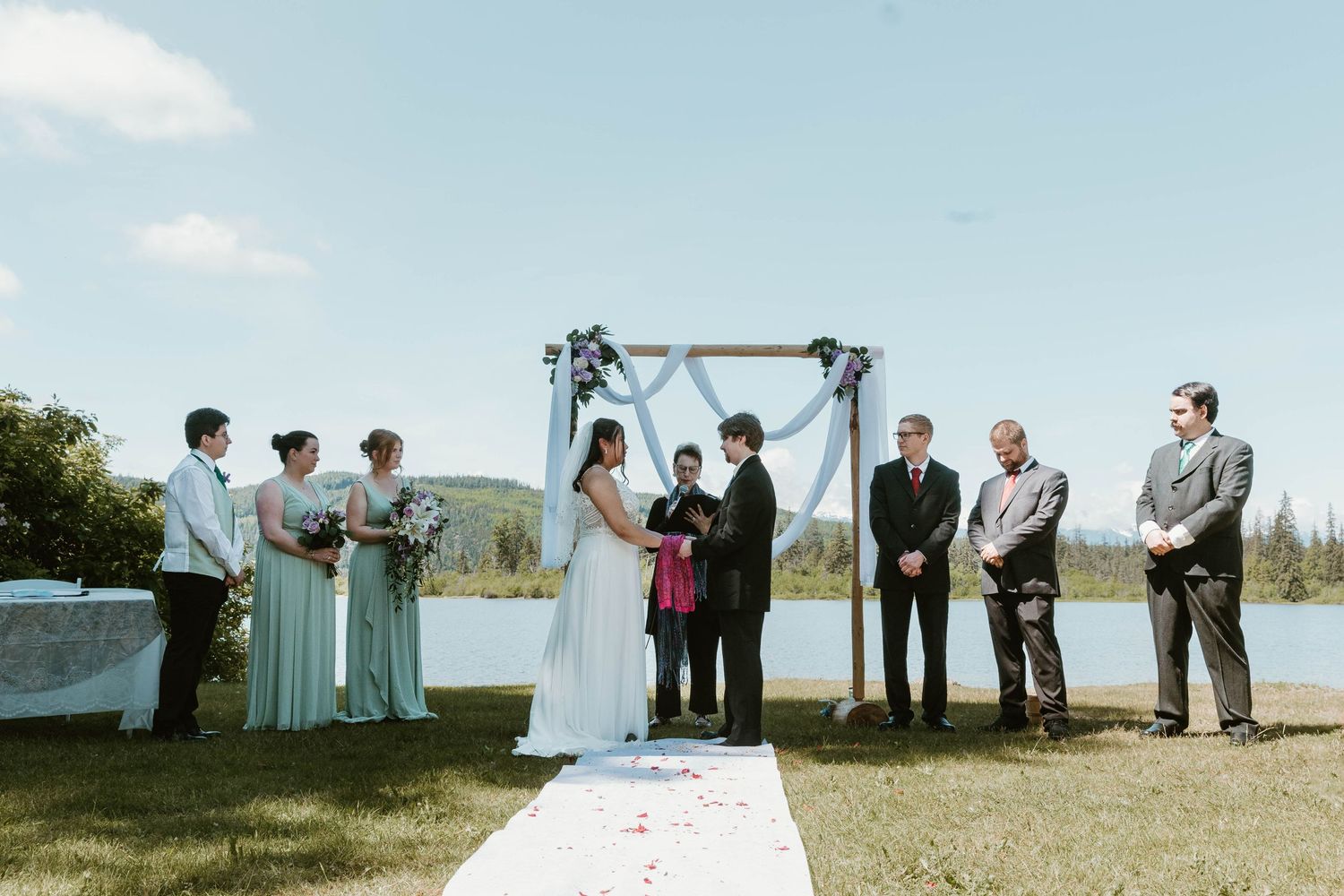 Wedding ceremony under an arch by a lake with wedding party lined up on both sides.