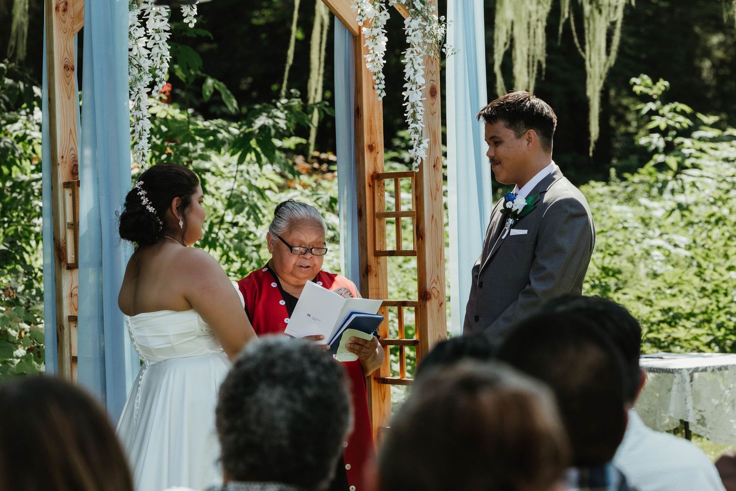 An outdoor wedding ceremony taking place under a wooden arch adorned with white flowers.