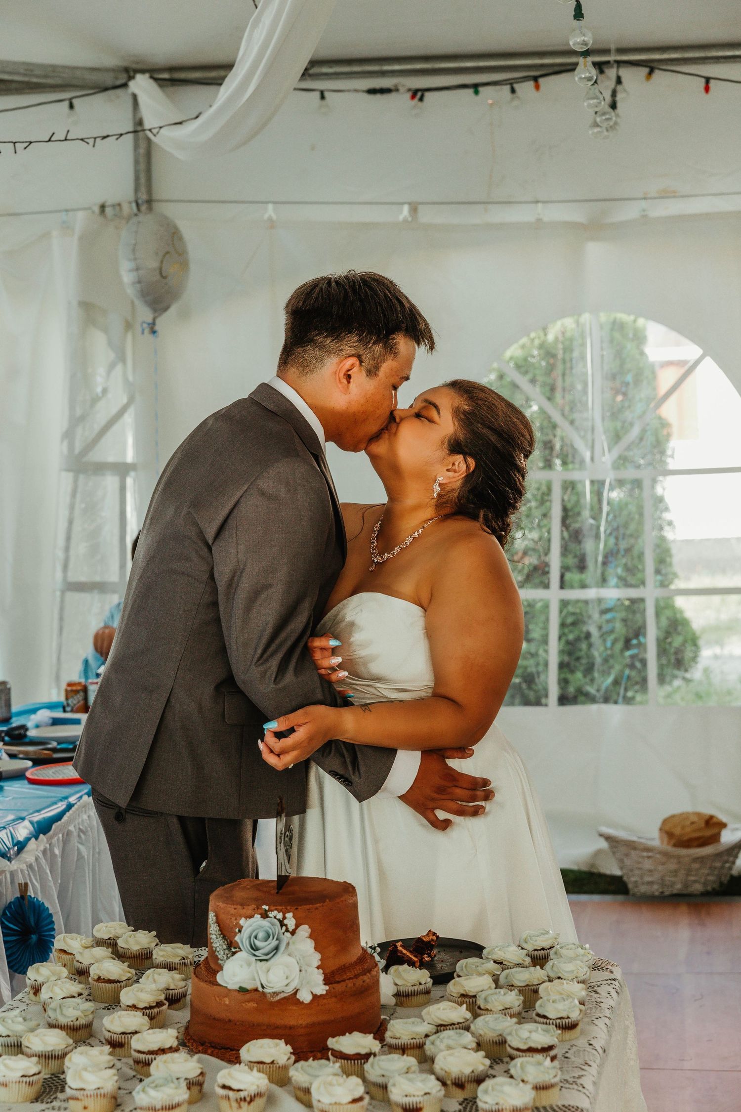 Newlyweds share a kiss by their cupcake display under a white tent.