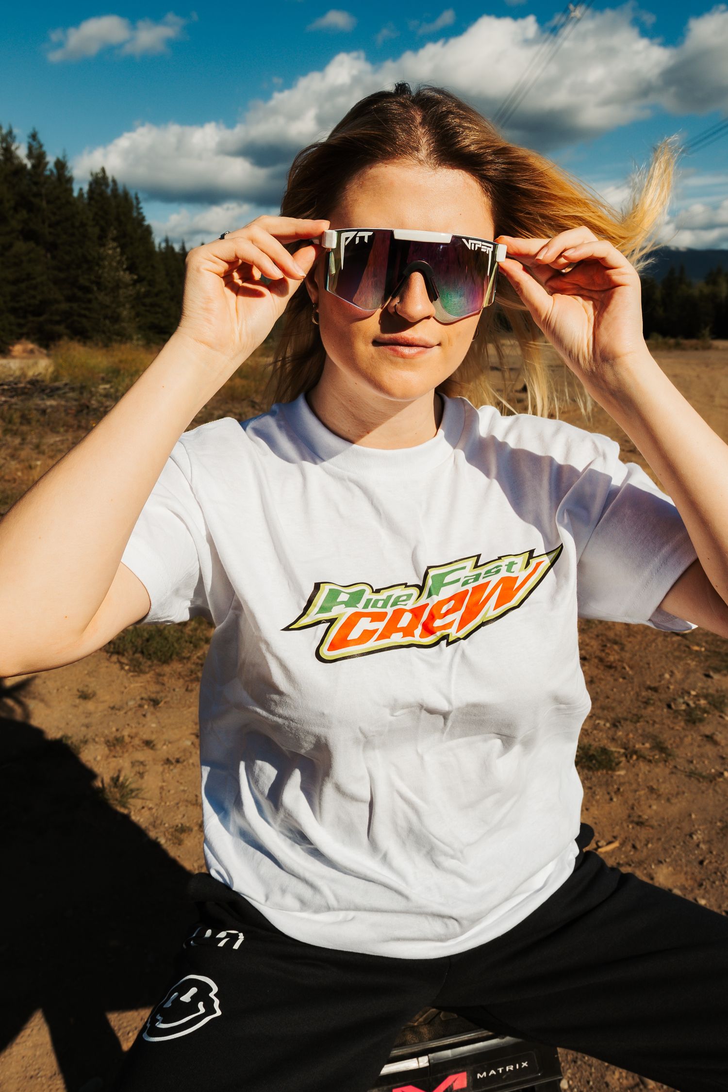 Person in white Mountain Dew t-shirt and sunglasses poses outdoors against blue sky and mountain landscape.