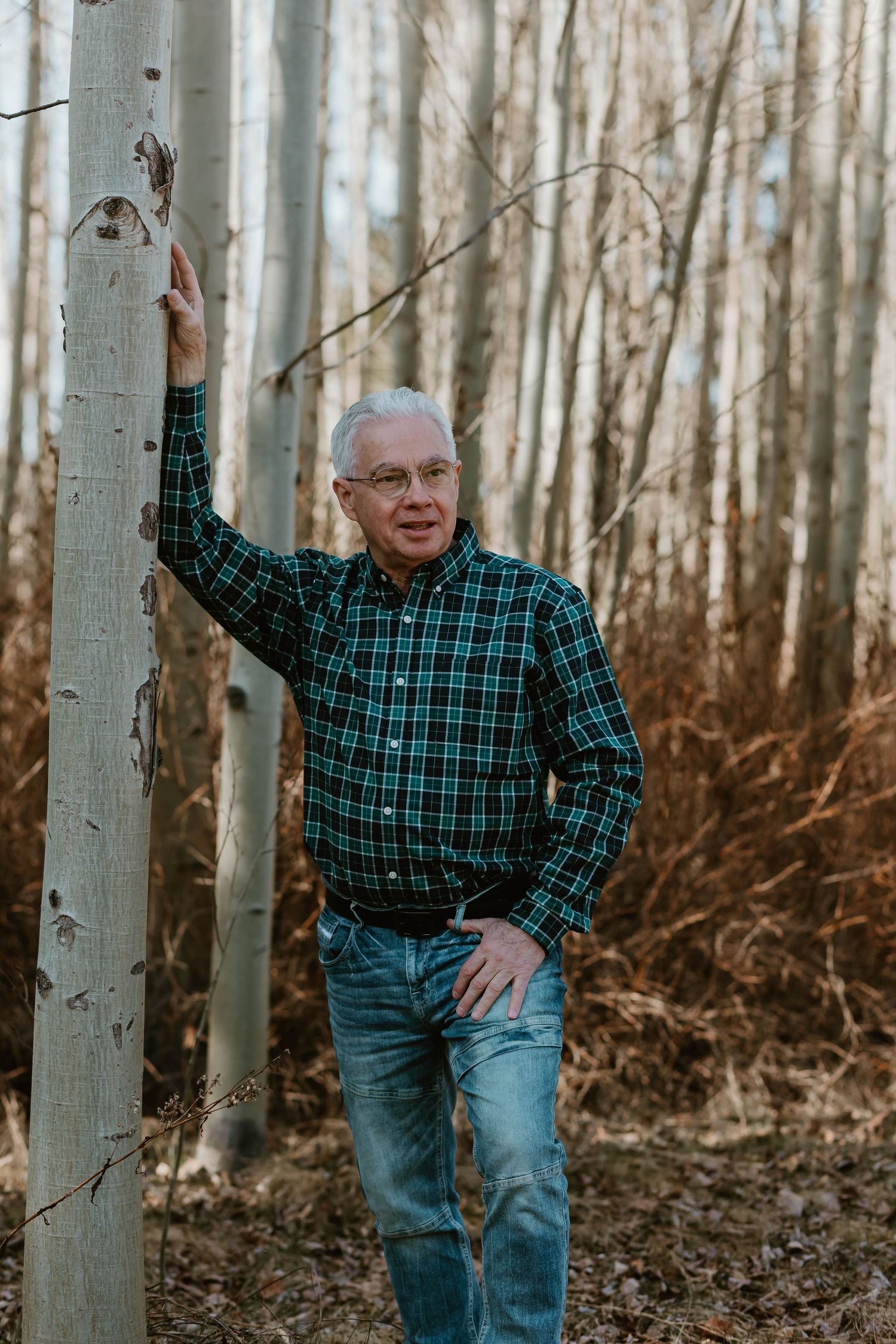 Person in a green plaid shirt and jeans stands casually against an aspen tree in a winter forest.