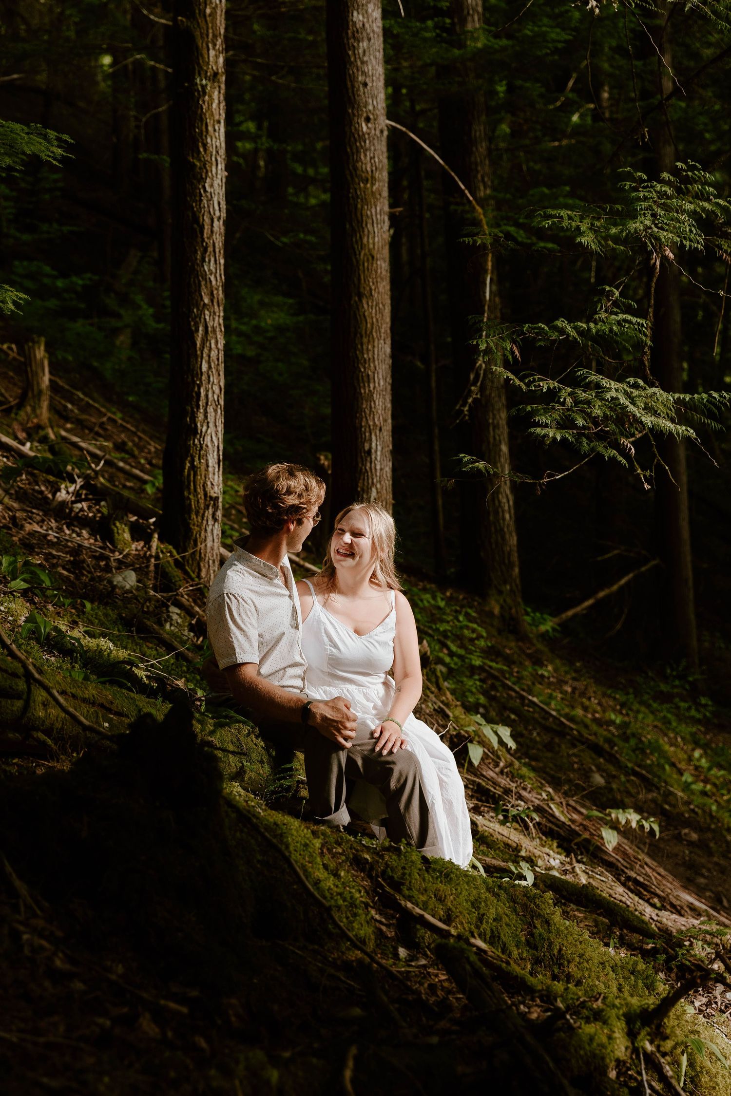 A joyful couple sitting on a moss-covered log in a lush forest setting.