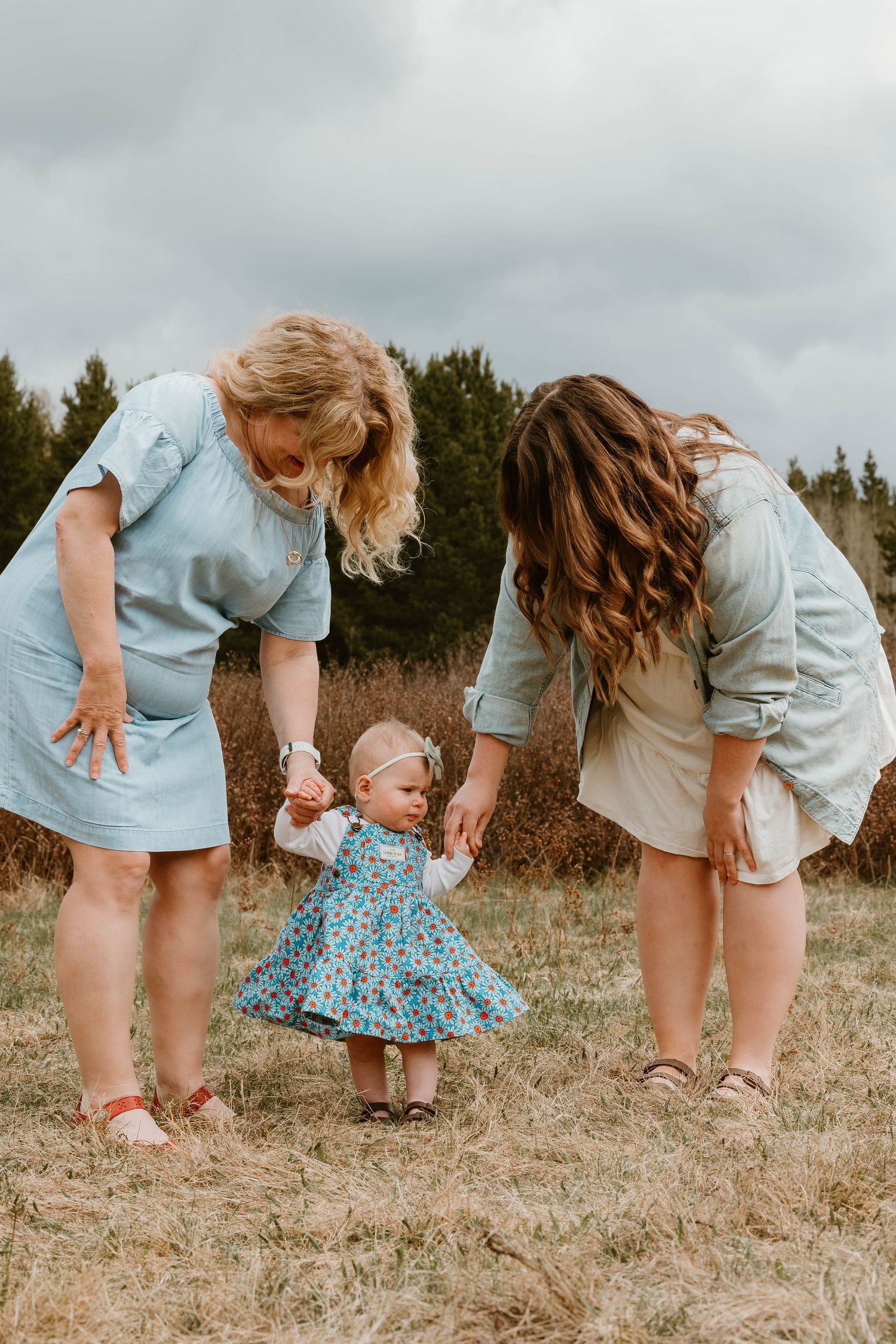 Three people in light blue dresses helping a small child take steps outdoors.