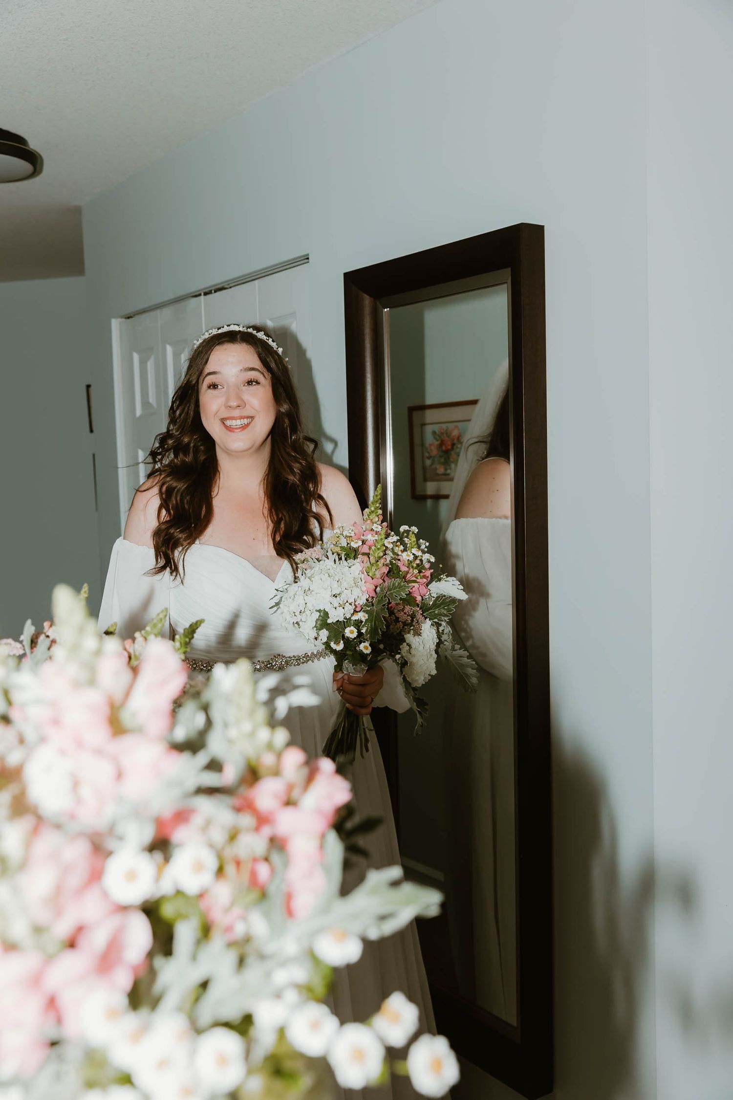 Bride in white dress holds bouquet while standing near mirror in natural light.