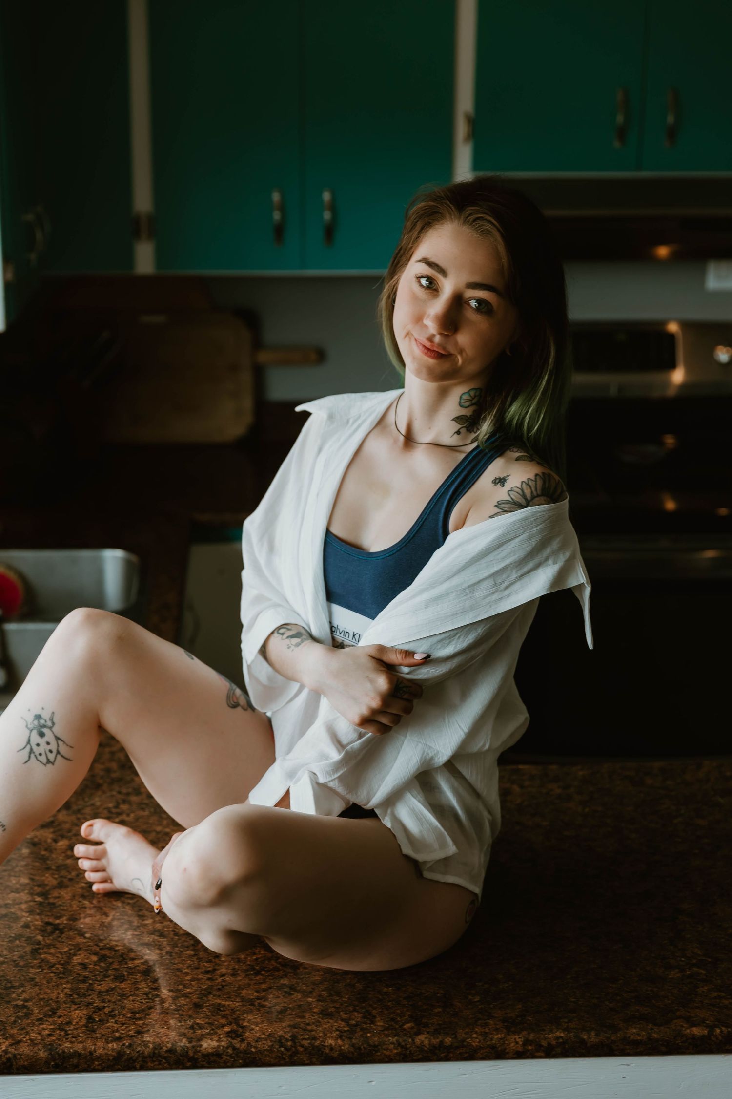 A person in a white button-down shirt and dark tank top poses on a kitchen counter in moody lighting.
