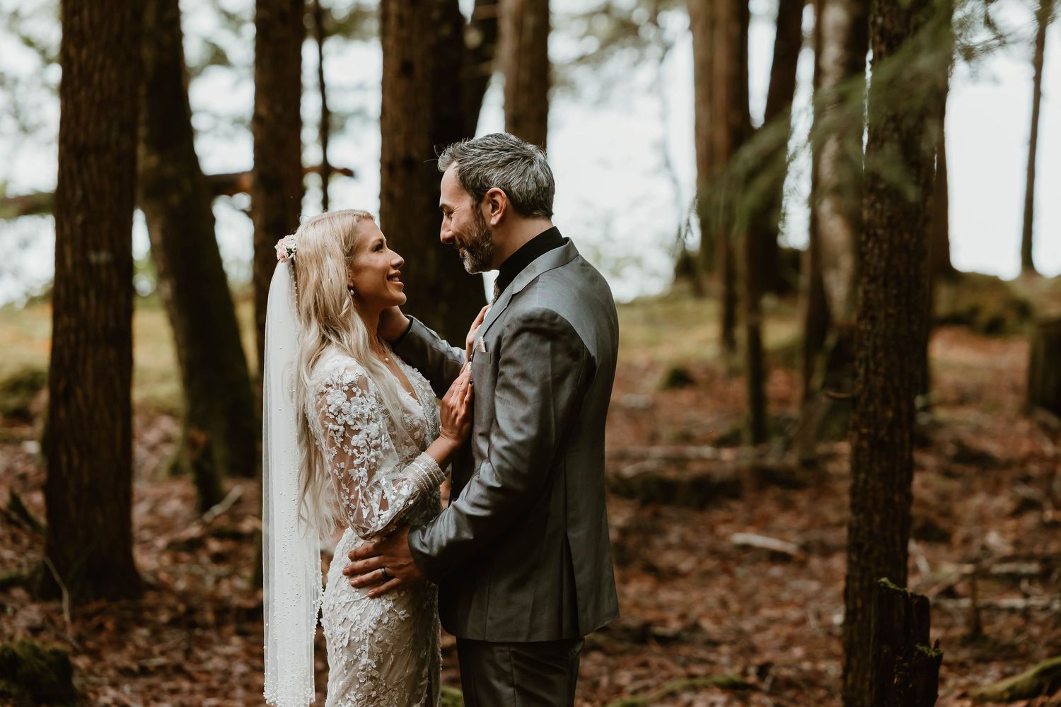 A couple embraces in a moody forest setting wearing elegant wedding attire among tall pine trees.