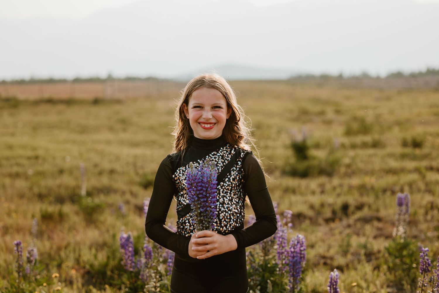 Person wearing black sweater stands in a field of purple wildflowers during golden hour at sunset.