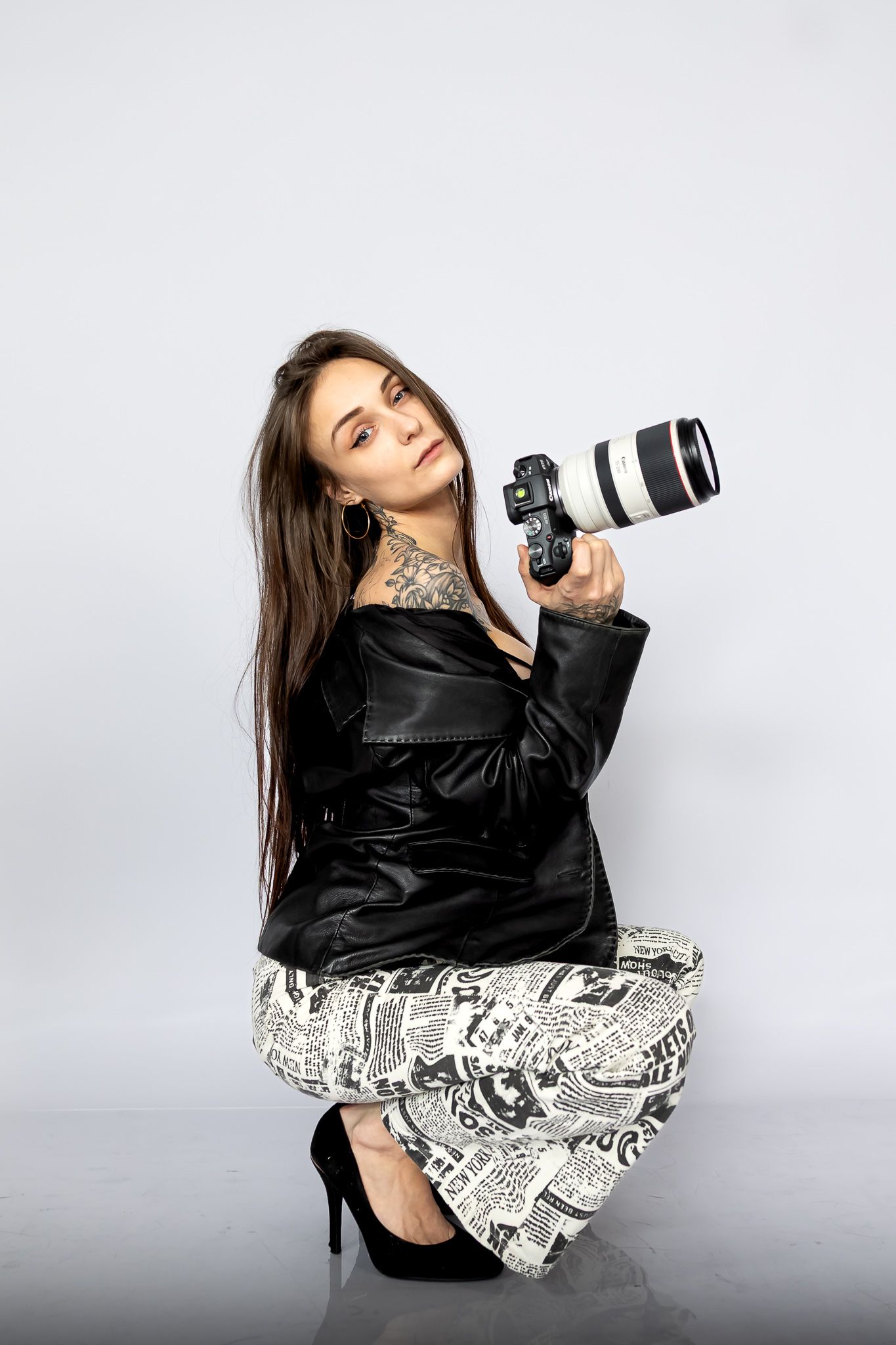 Professional photographer in black leather jacket and patterned dress poses with DSLR camera and telephoto lens against gray background.