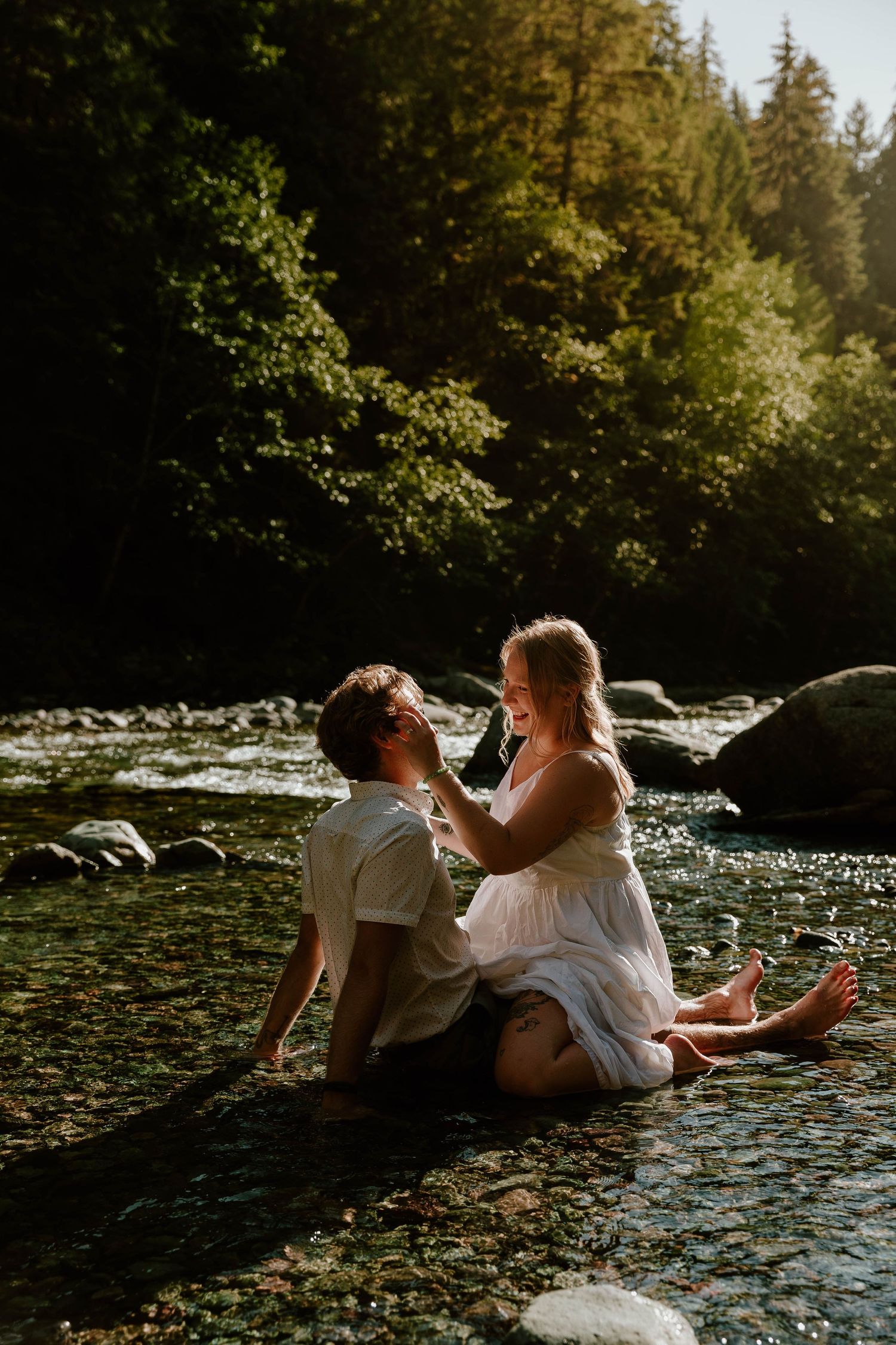 Couple laughs together while sitting in shallow river water during romantic photoshoot.