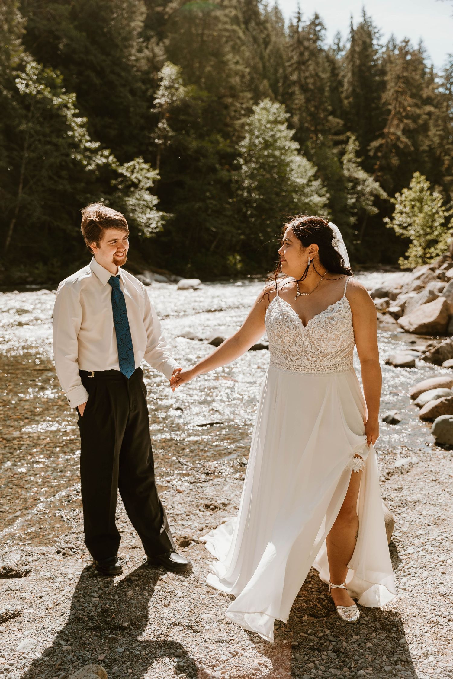 A couple shares a joyful moment on a rocky riverbank surrounded by evergreen trees.