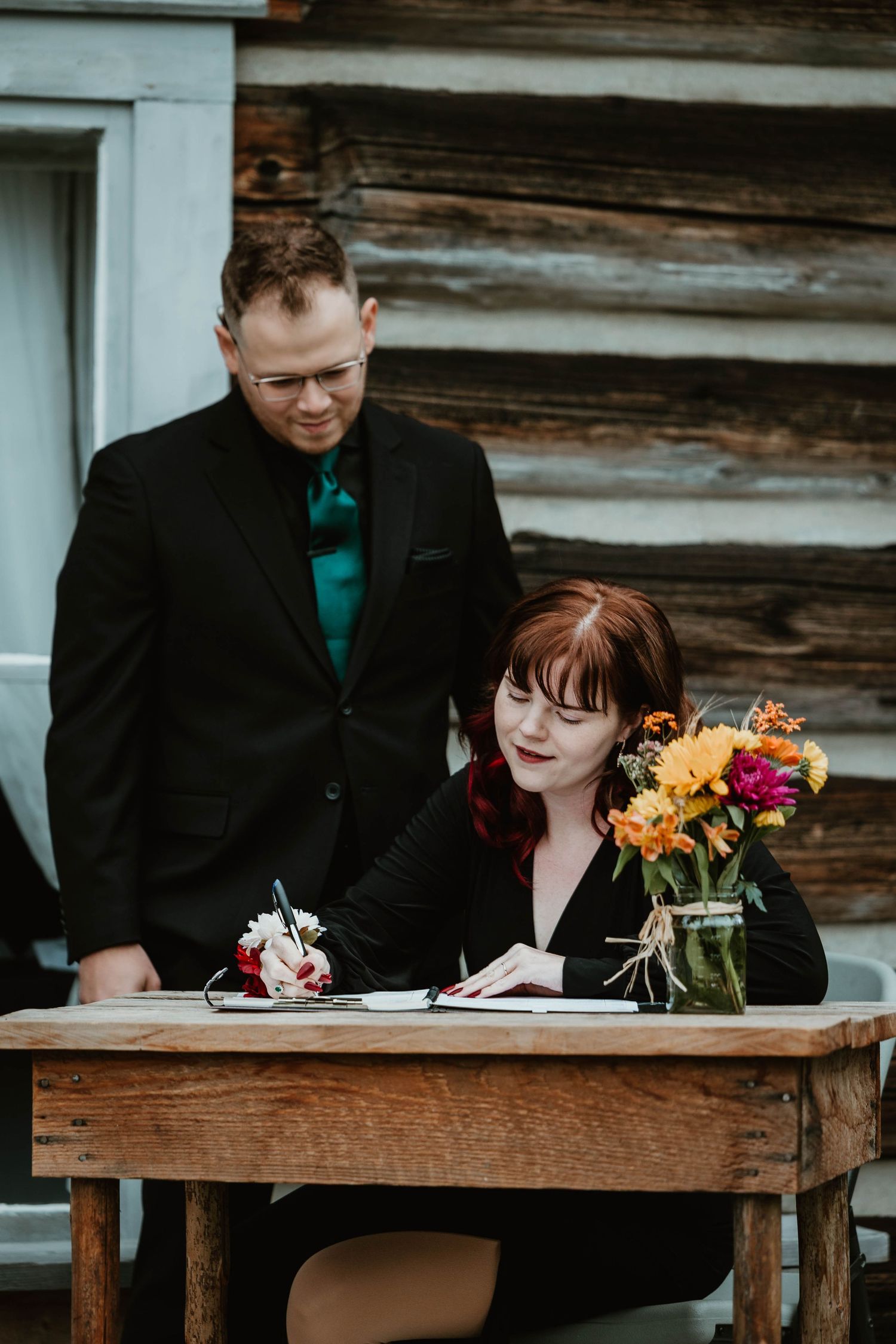 A couple signs documents at a rustic wooden table with colorful flowers.