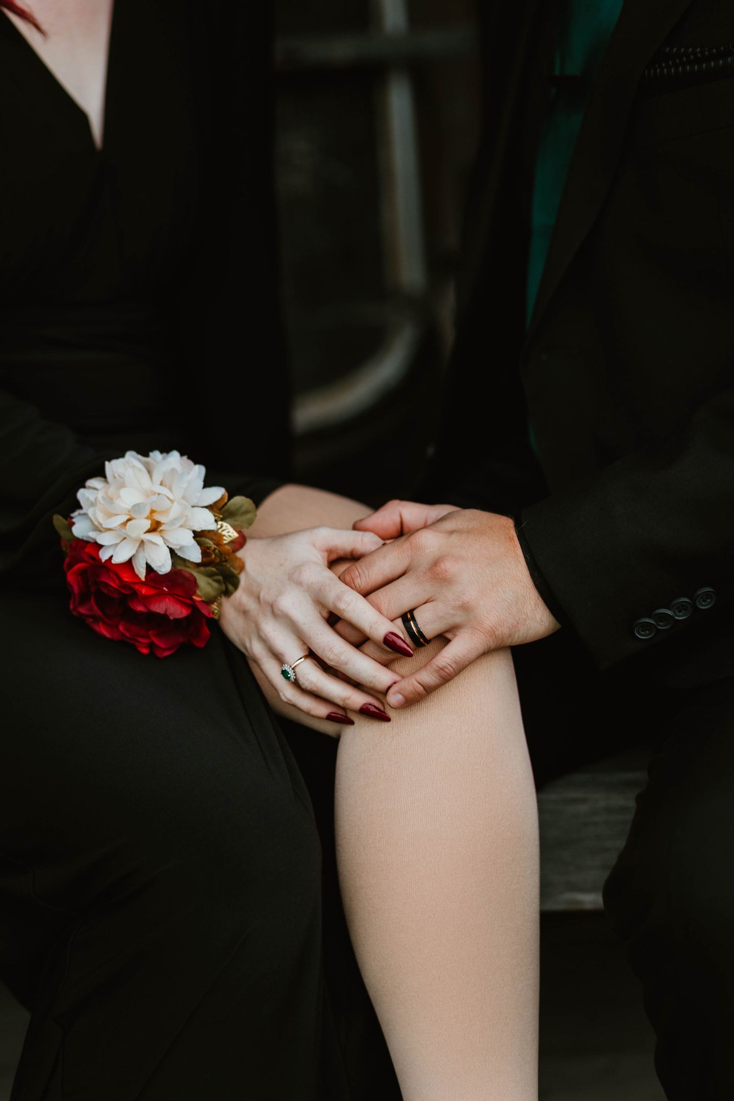 Hands with red nail polish intertwined showing rings and a floral corsage against formal black attire.