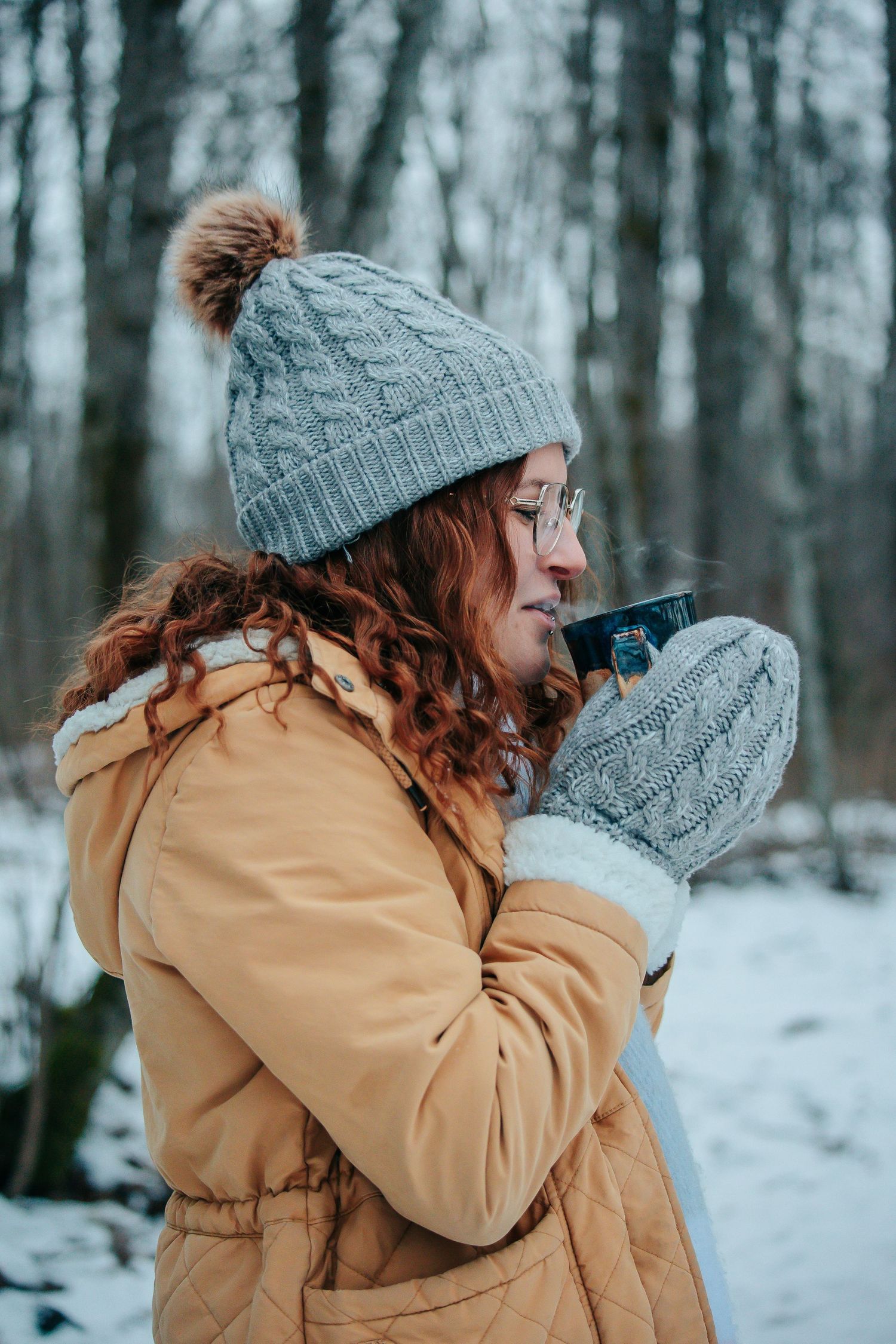 Person in cozy winter attire sips from a mug in a snowy forest setting.