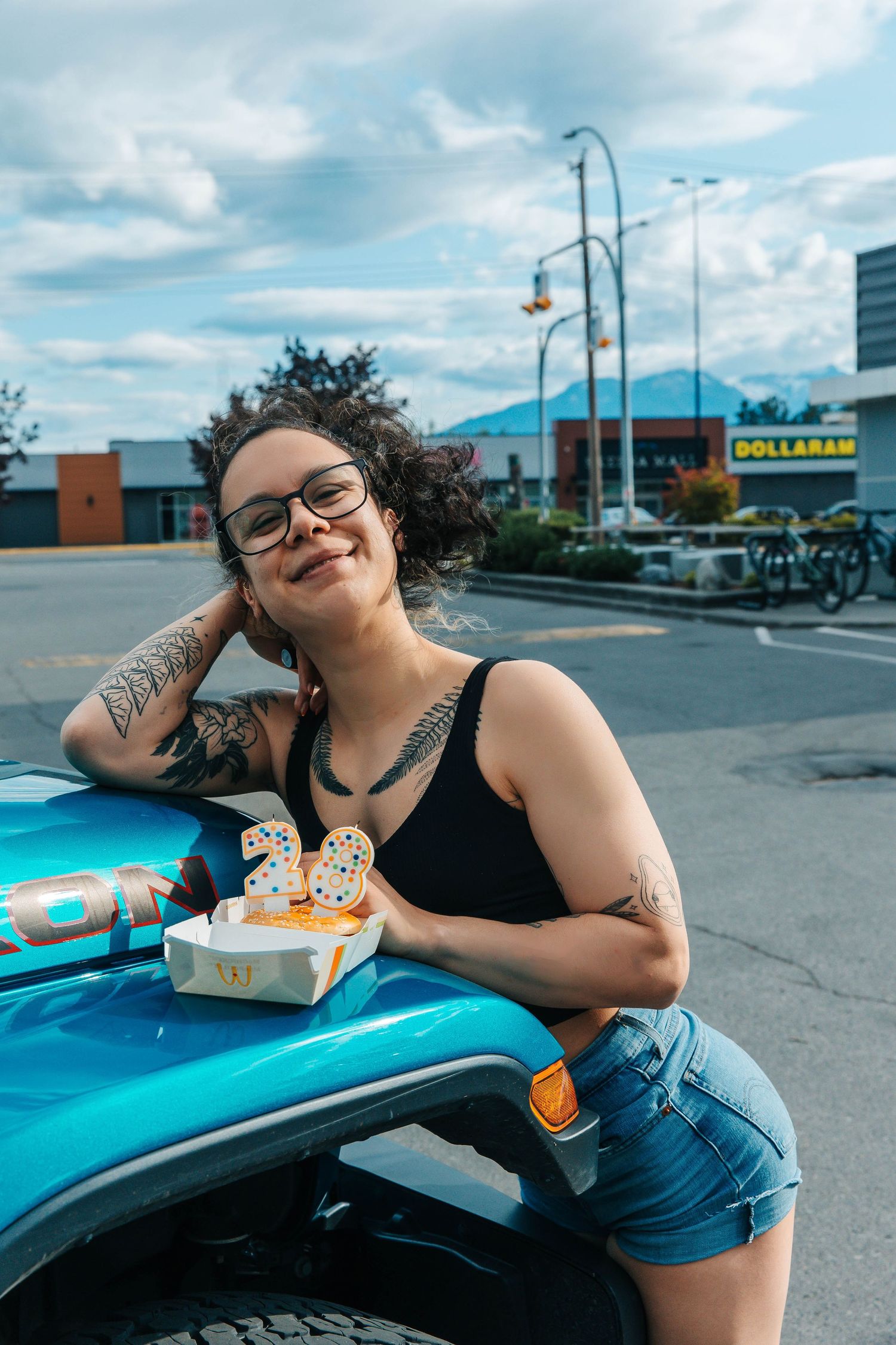 A casual photo in a parking lot with blue car and glasses, showing tattooed arm and bright smile.