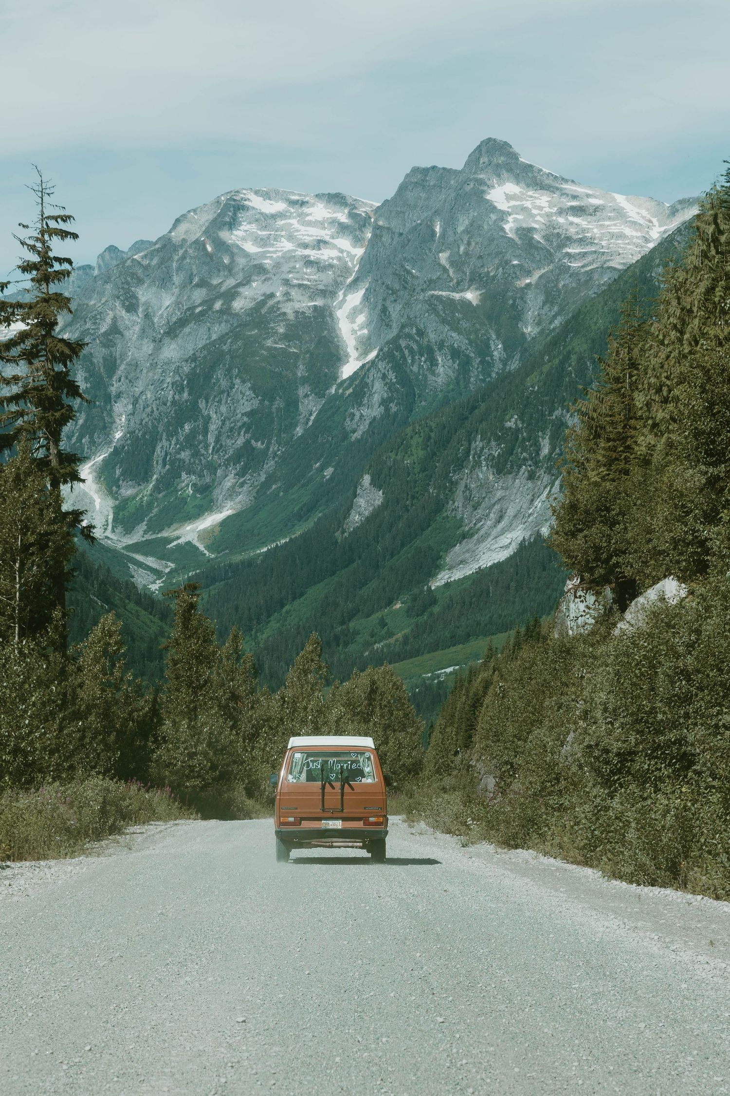 An orange vintage van drives down a mountain road with snow-capped peaks in the distance.