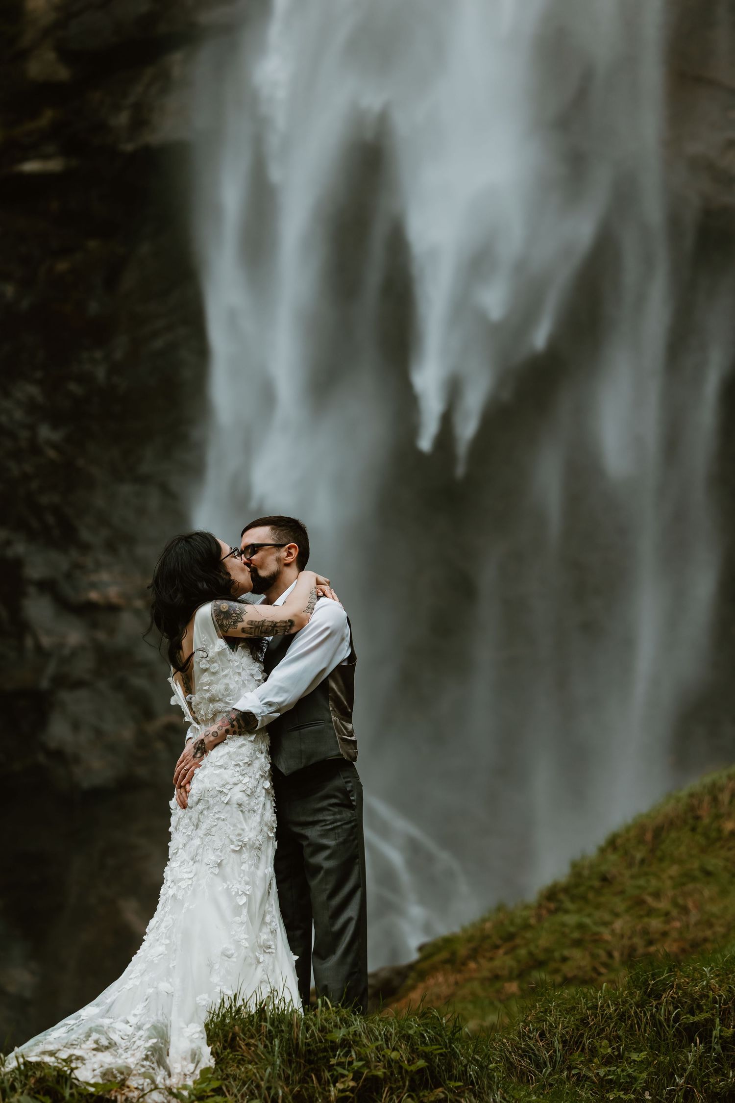 A couple shares a romantic embrace in front of a misty waterfall, the bride wearing a lace gown and groom in a vest.