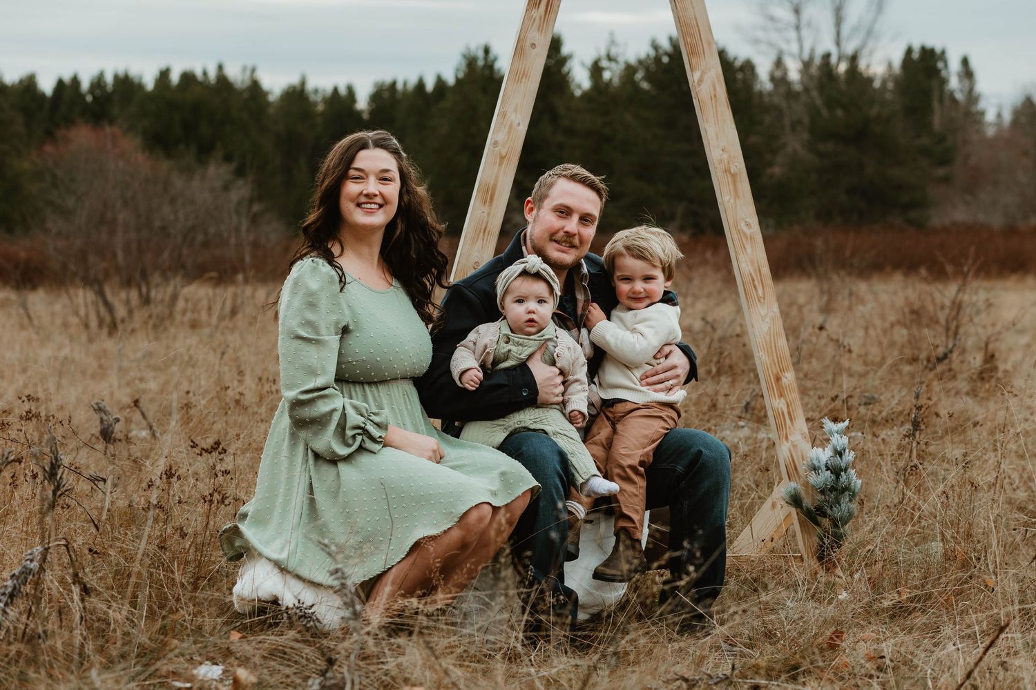 A family sits together on a rustic wooden swing in a golden field during autumn with evergreen trees in the background.