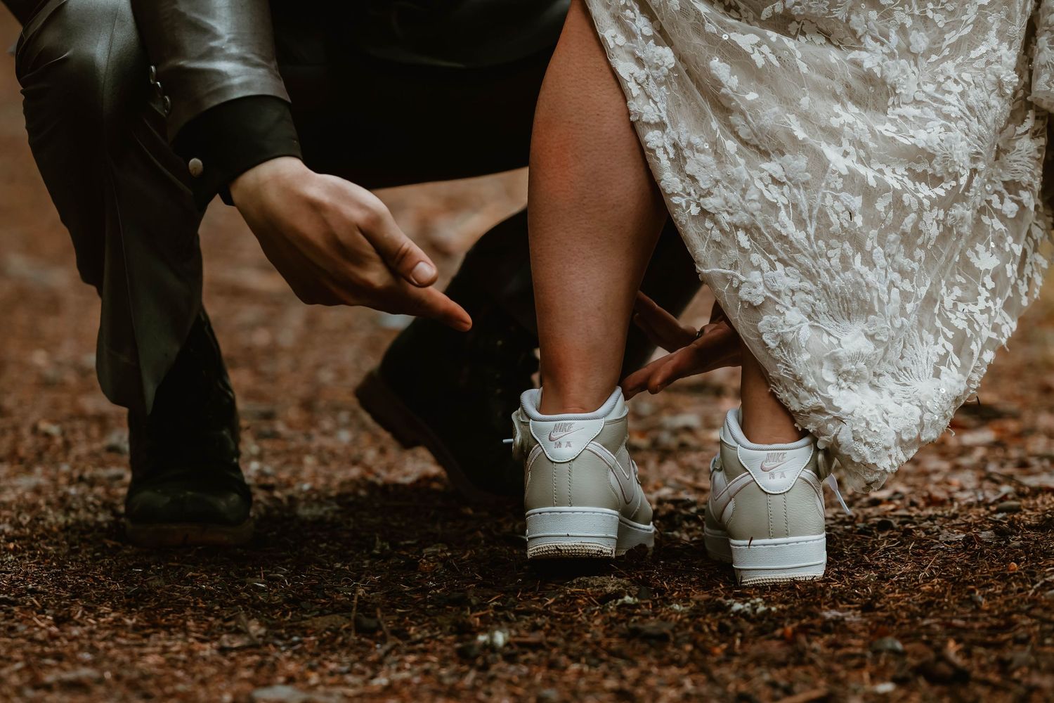 Close-up shot of bride in white wedding dress and sneakers alongside groom's feet on brown textured ground.