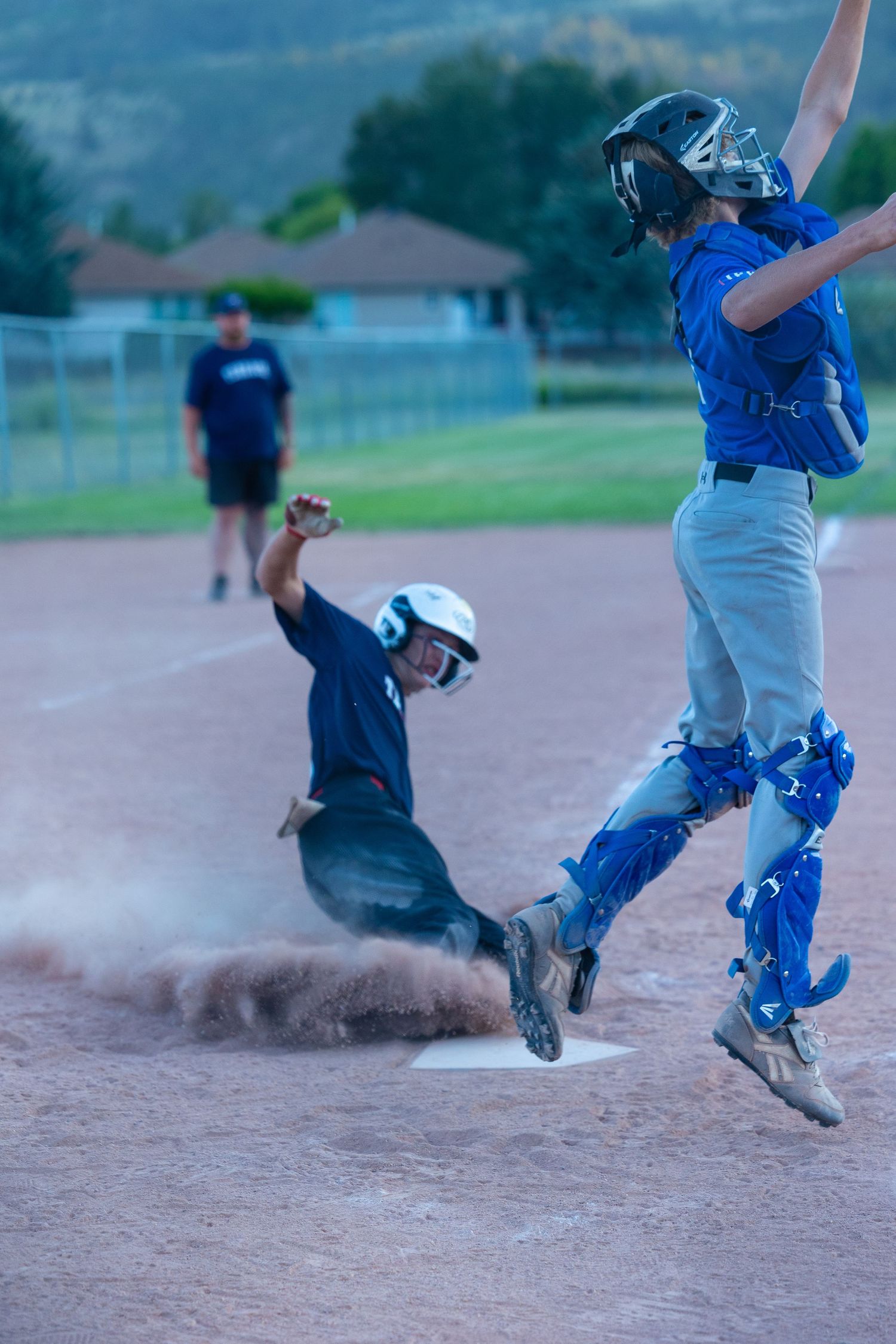 Dynamic baseball action shot showing players jumping and sliding at home plate with dust flying.