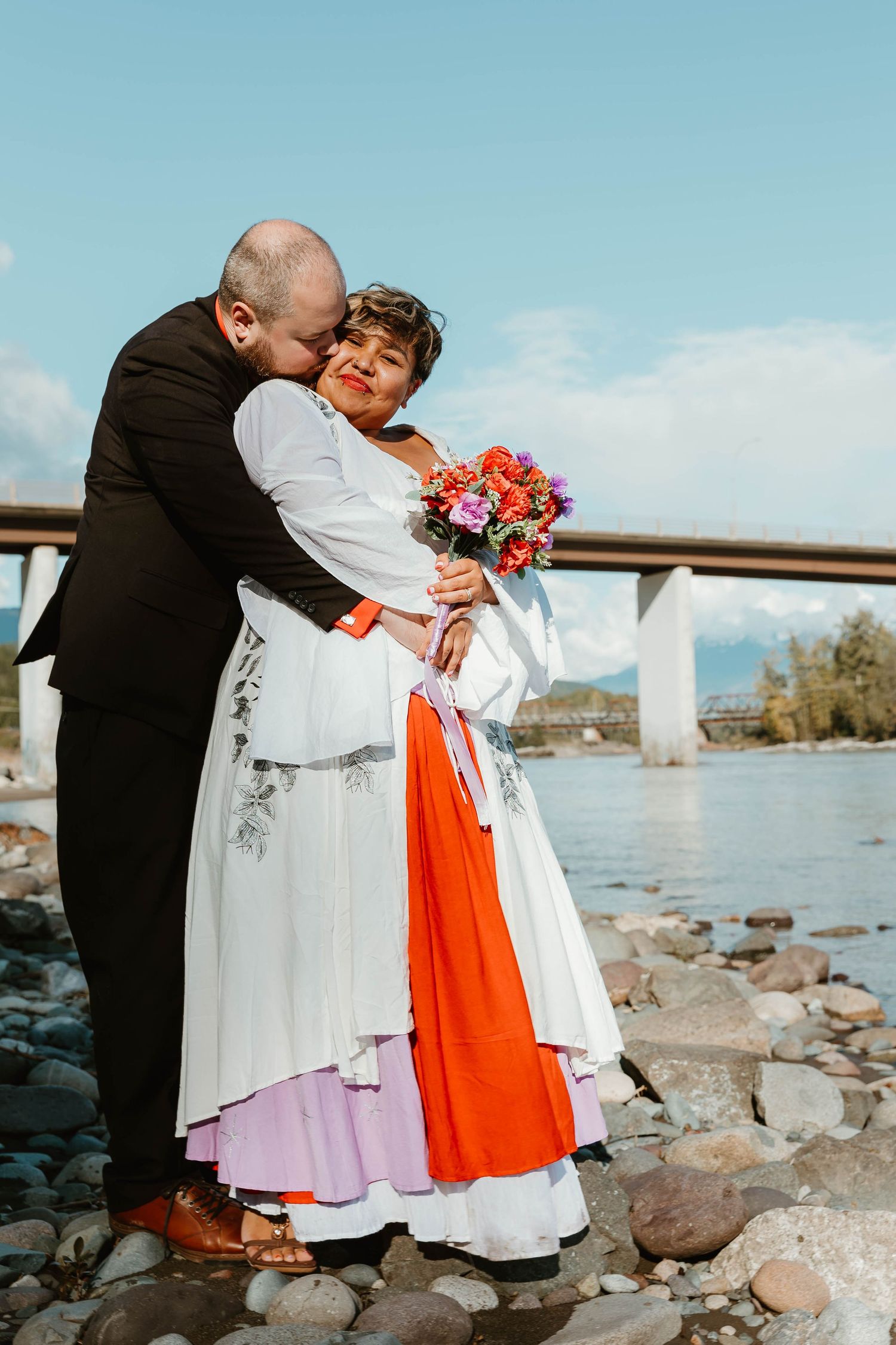 A couple shares an intimate moment by a river while dressed in wedding attire with orange and white details.