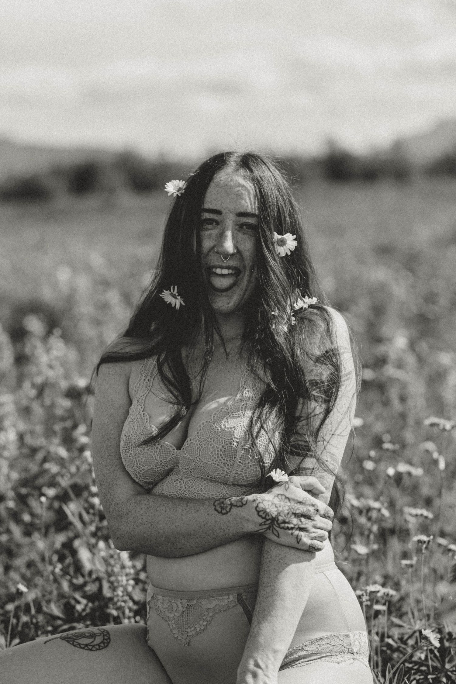 Black and white photo of someone sitting in a flower field wearing a sweater with daisies in their hair.