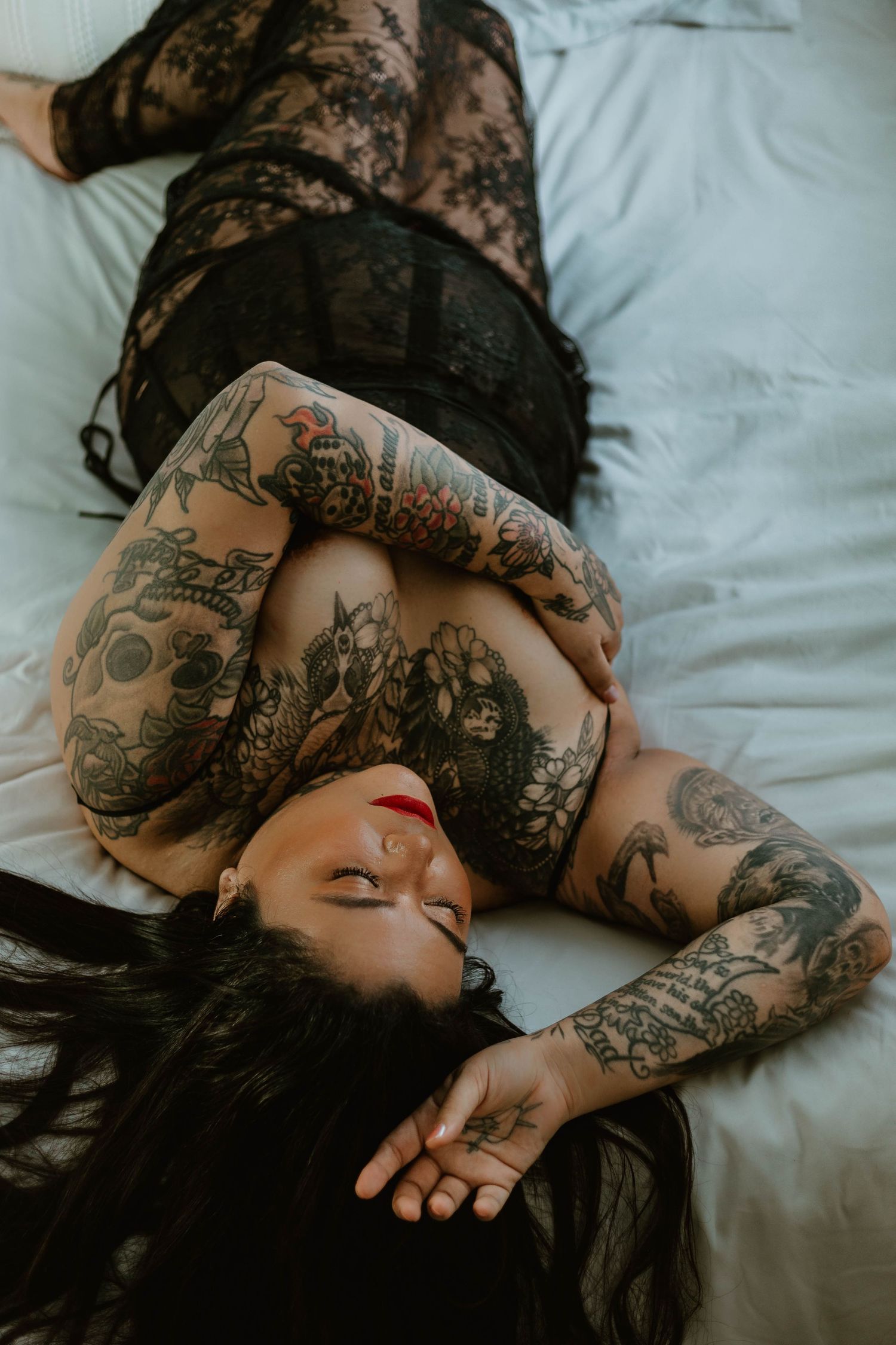 Artistic boudoir shot featuring black lace and extensive tattoo artwork on white sheets.