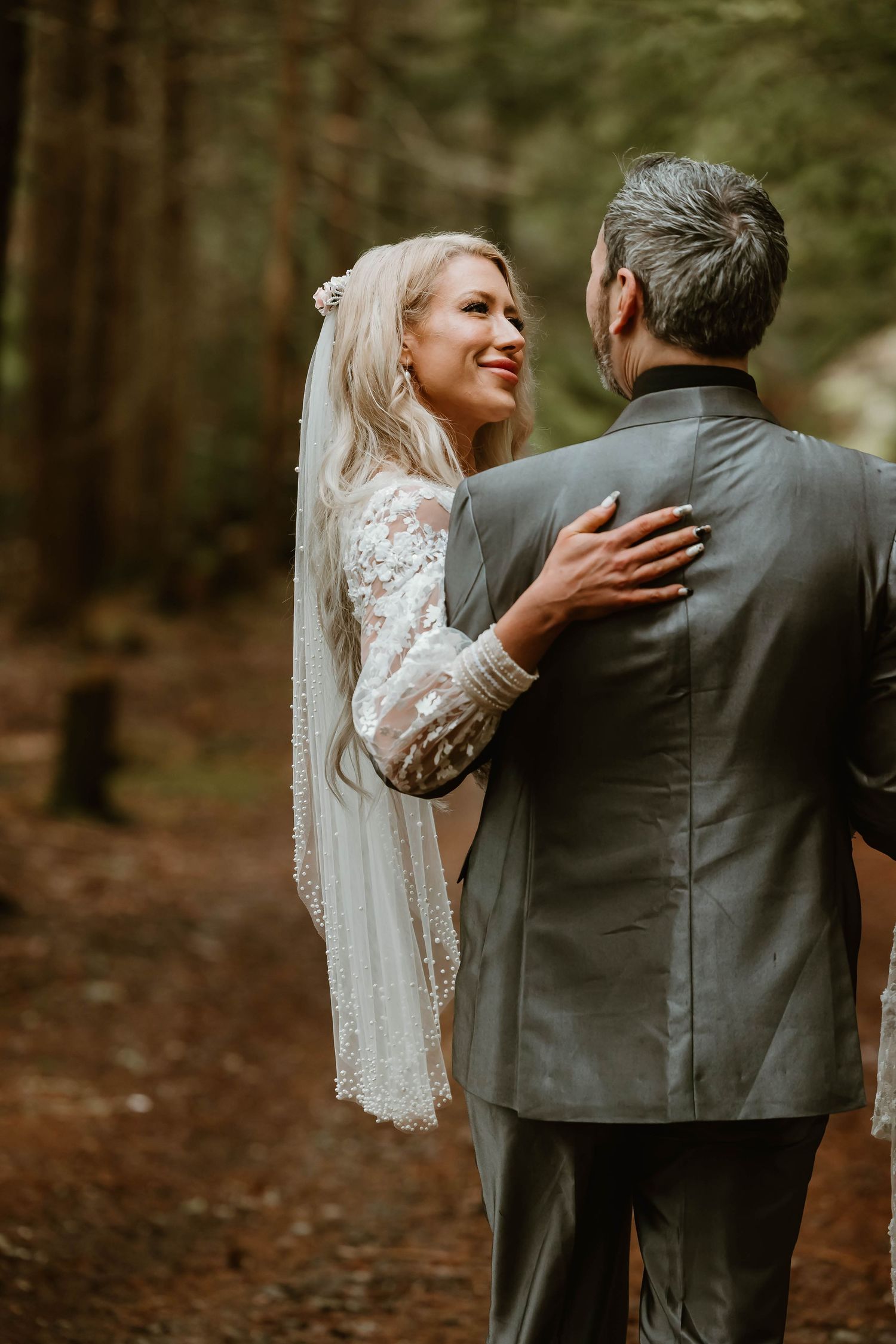 A romantic outdoor wedding photo of a bride in lace embracing her groom in a forest setting.
