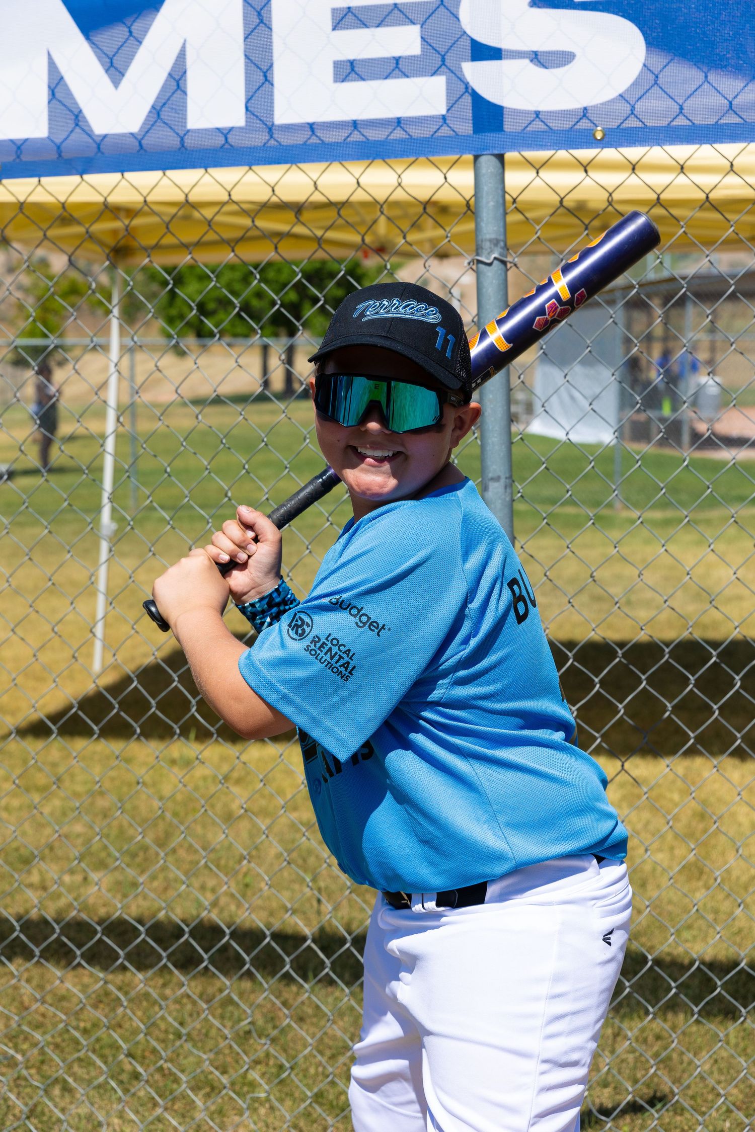 Youth baseball player in blue uniform posing with bat against chain link fence.