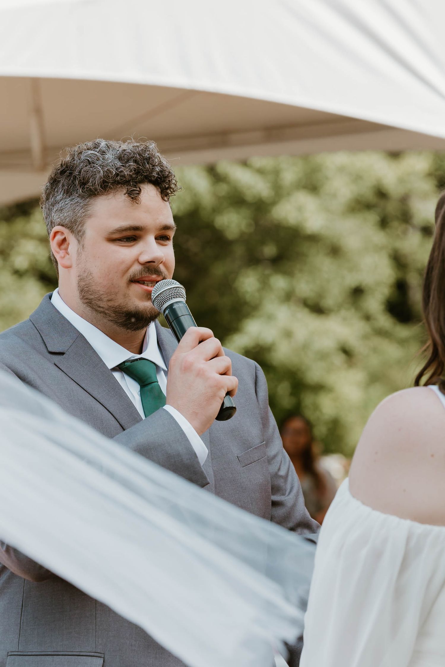 Groom in grey suit with green tie speaks into microphone during outdoor wedding ceremony.