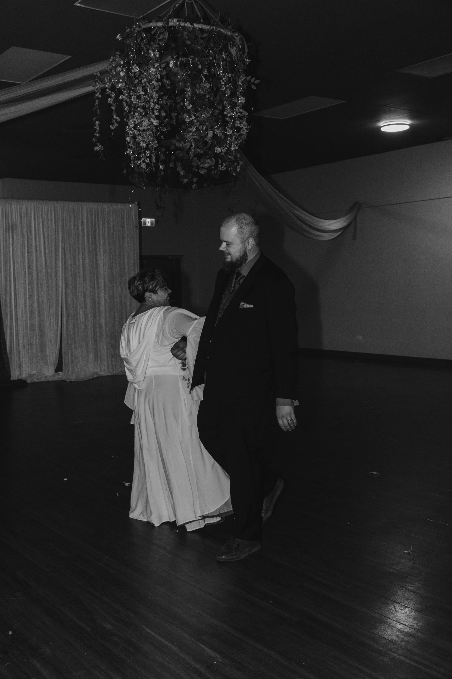 A black and white photo of a couple dancing under decorative hanging lights at an indoor venue.