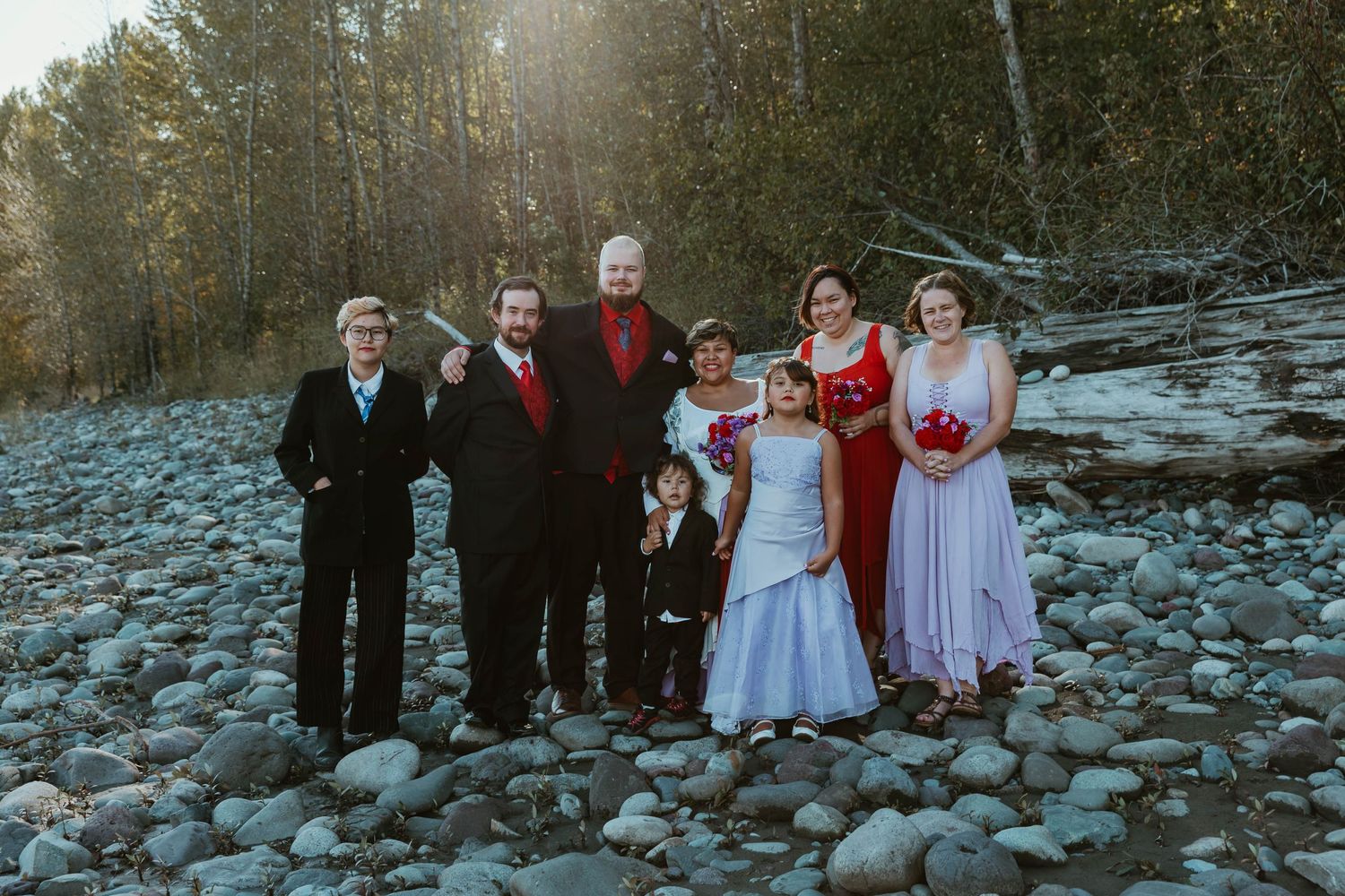 Wedding party poses for formal photo on rocky riverbank with forest backdrop.