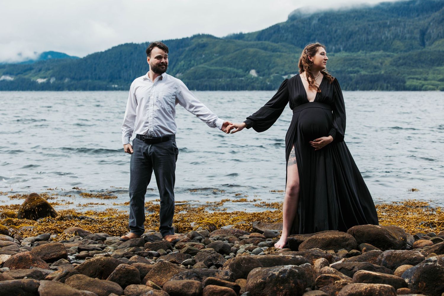 A couple holds hands while standing on rocky beach with misty mountains in background.