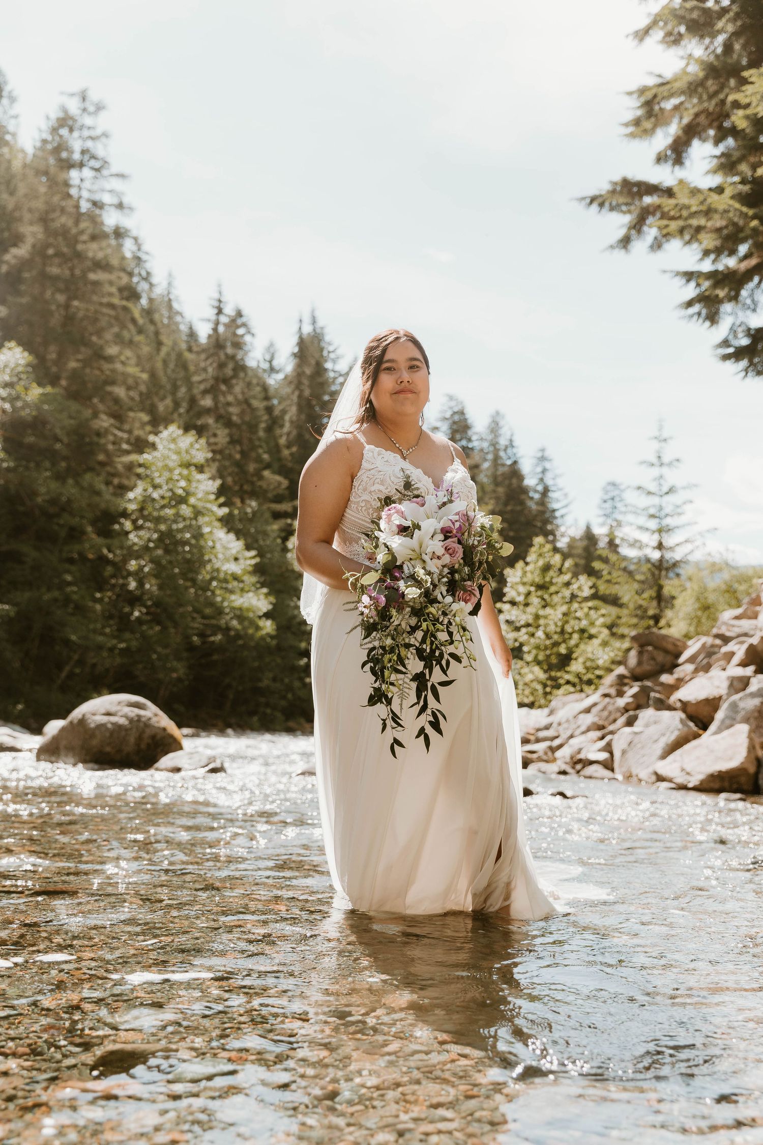 Person in white wedding dress stands in mountain stream holding cascading floral bouquet.