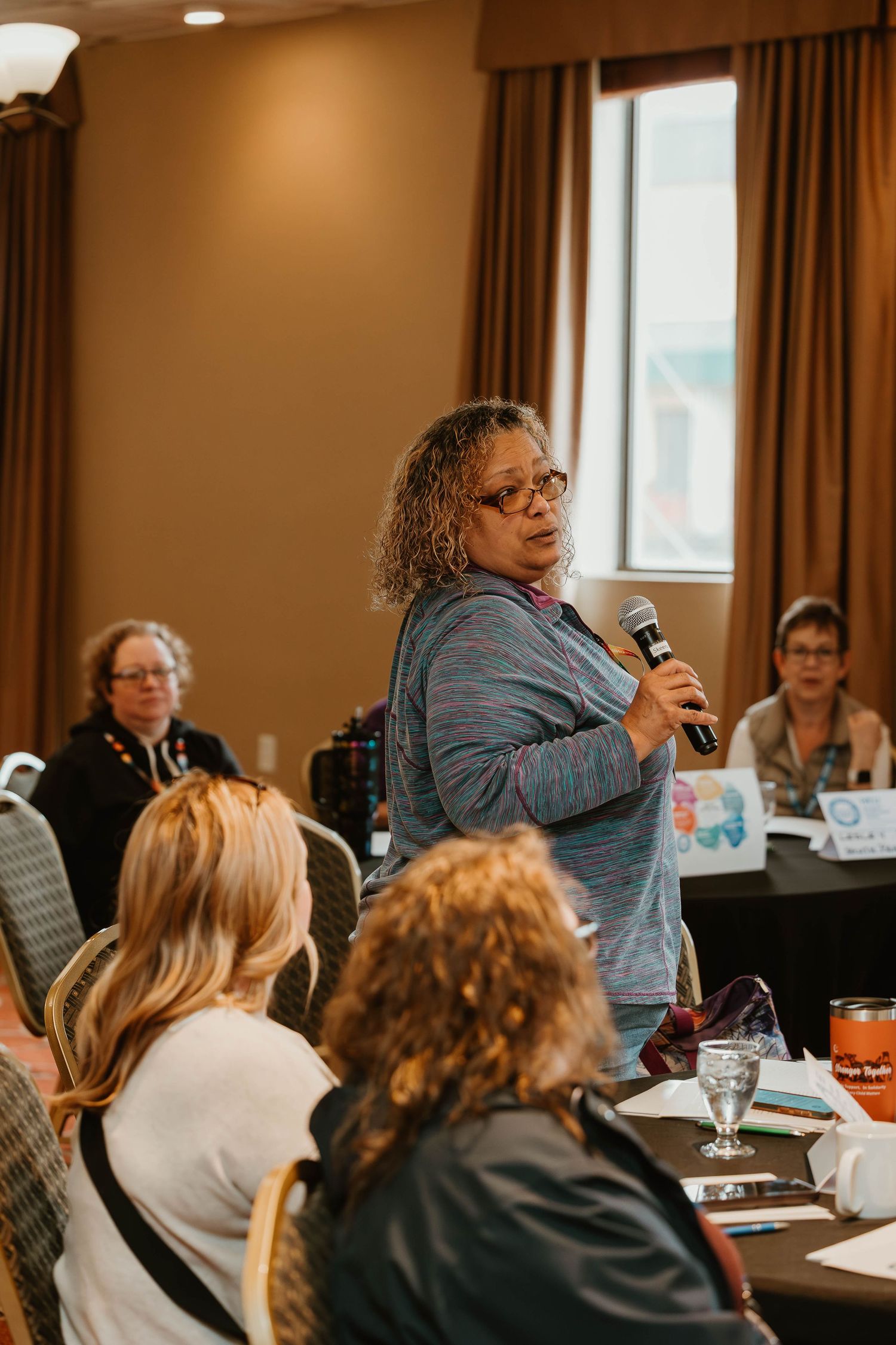 A speaker addresses the audience during a workshop session in a hotel conference room with brown curtains.