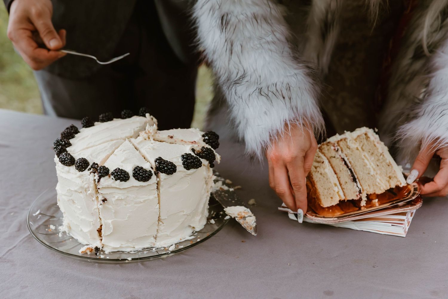 Two hands serving slices of a white frosted cake decorated with dark berries and cream filling.