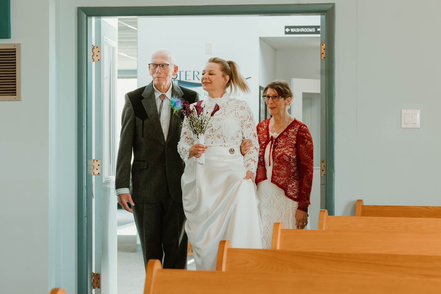 A bride in a white lace dress enters through blue church doors accompanied by attendants.
