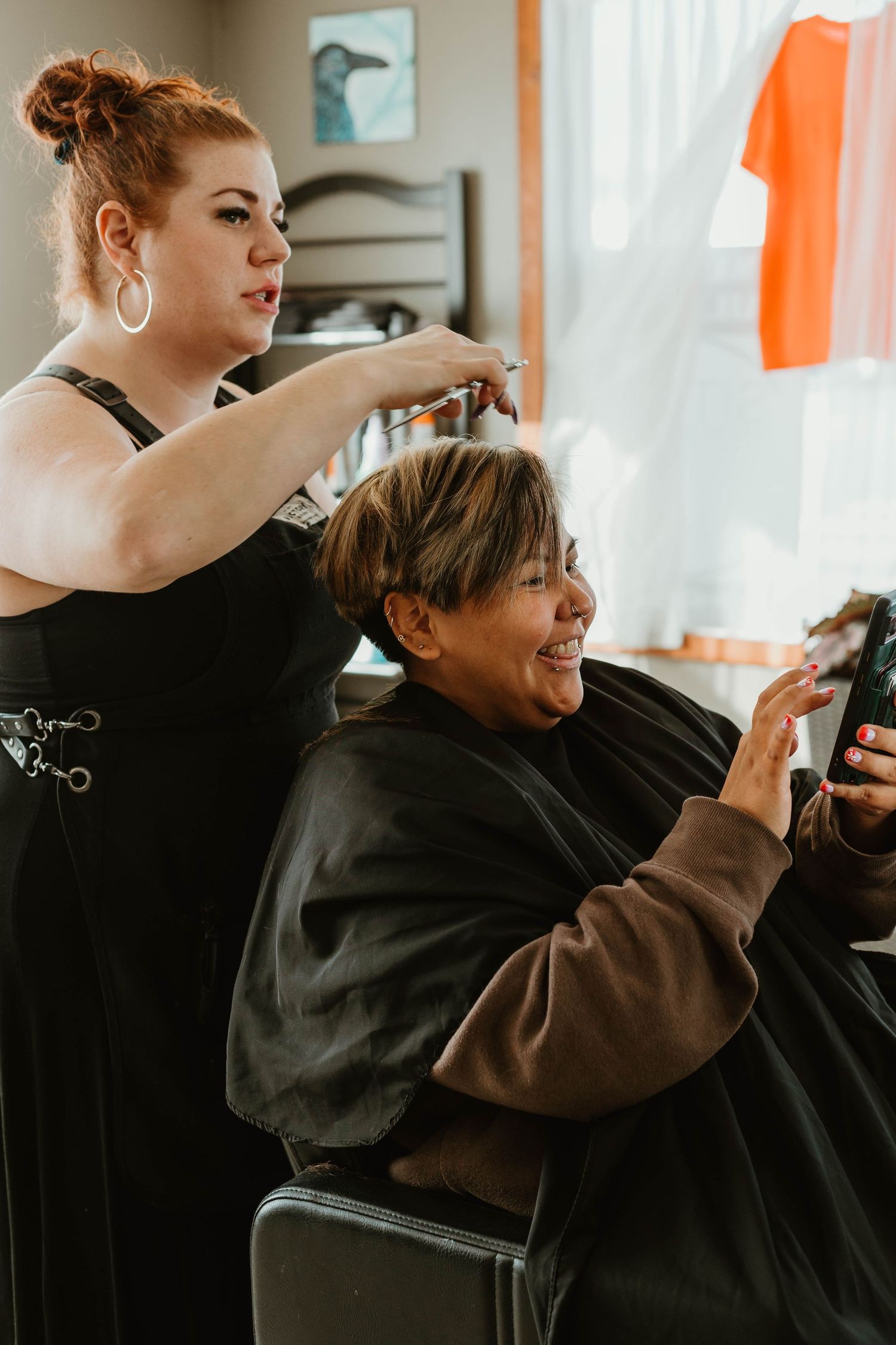 Stylist works on client's hair while client smiles and looks at phone in salon chair.