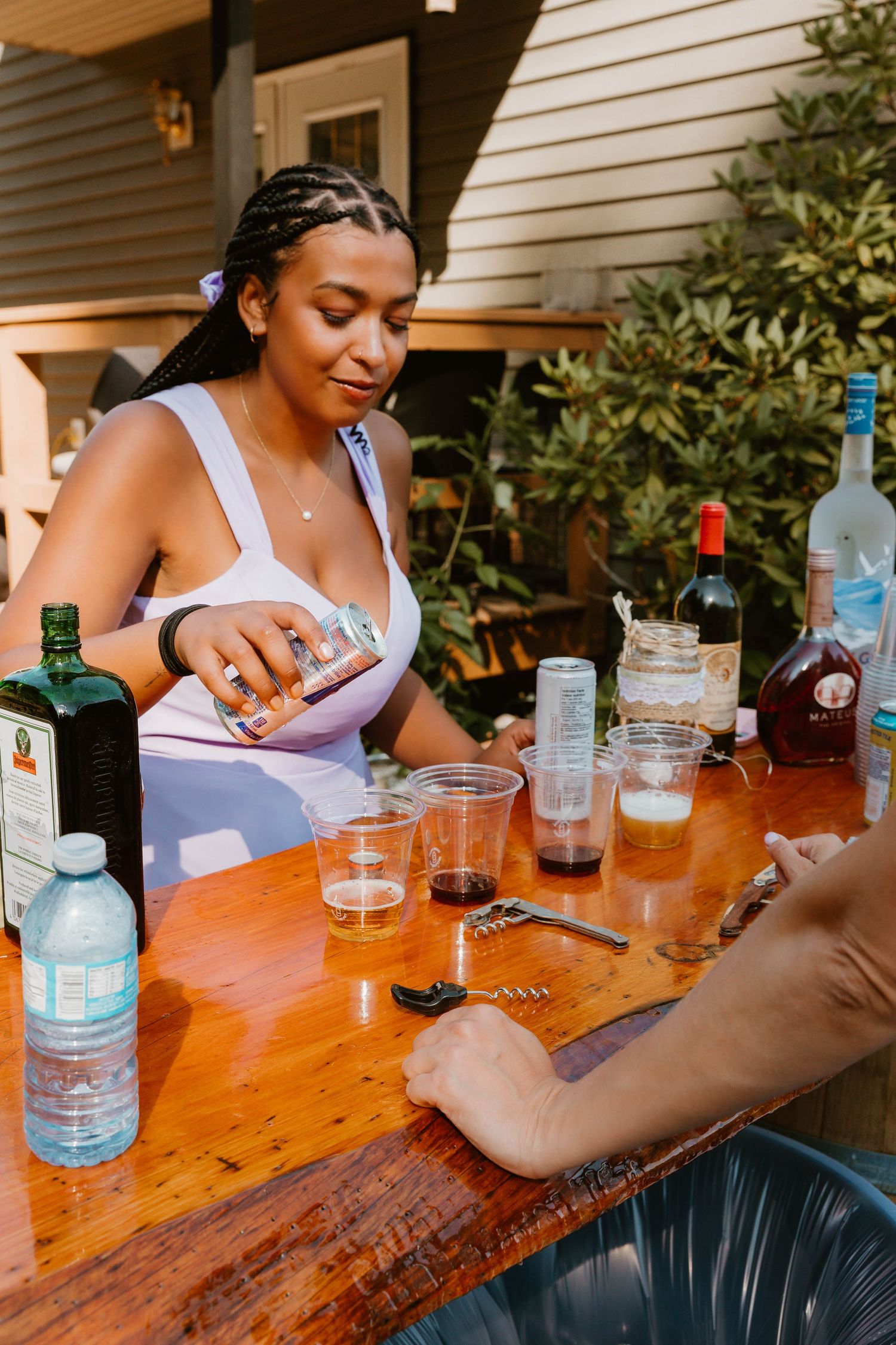 Person pouring drinks at an outdoor party with bottles and glasses on wooden table.