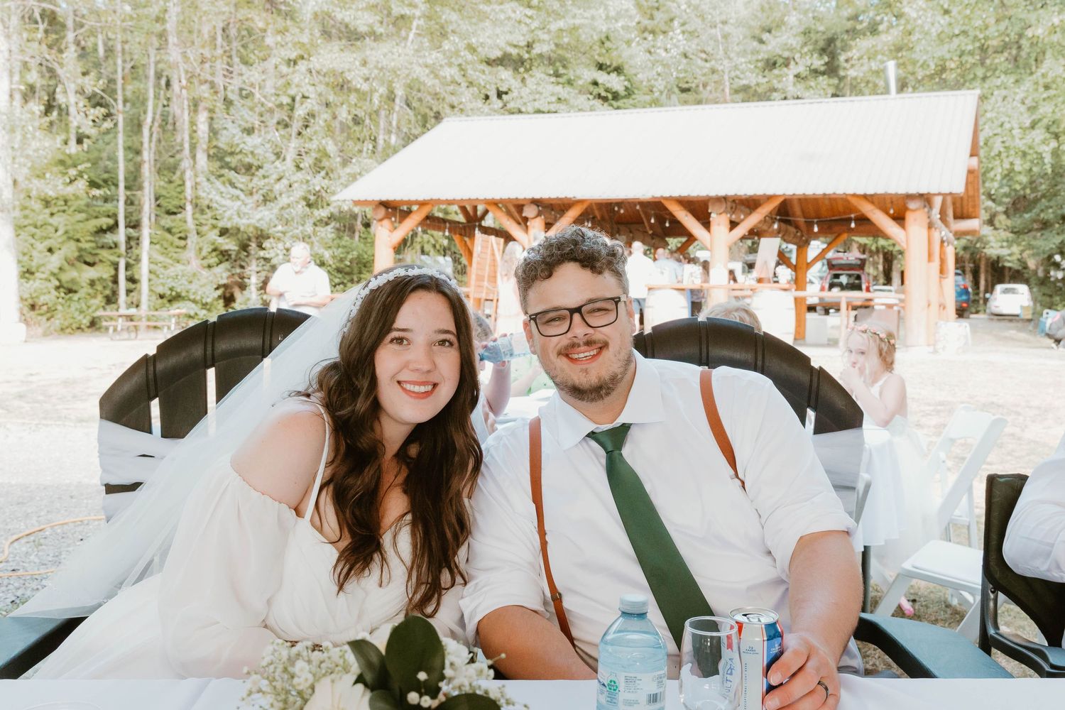 Newlyweds sitting together at outdoor reception under wooden pavilion.