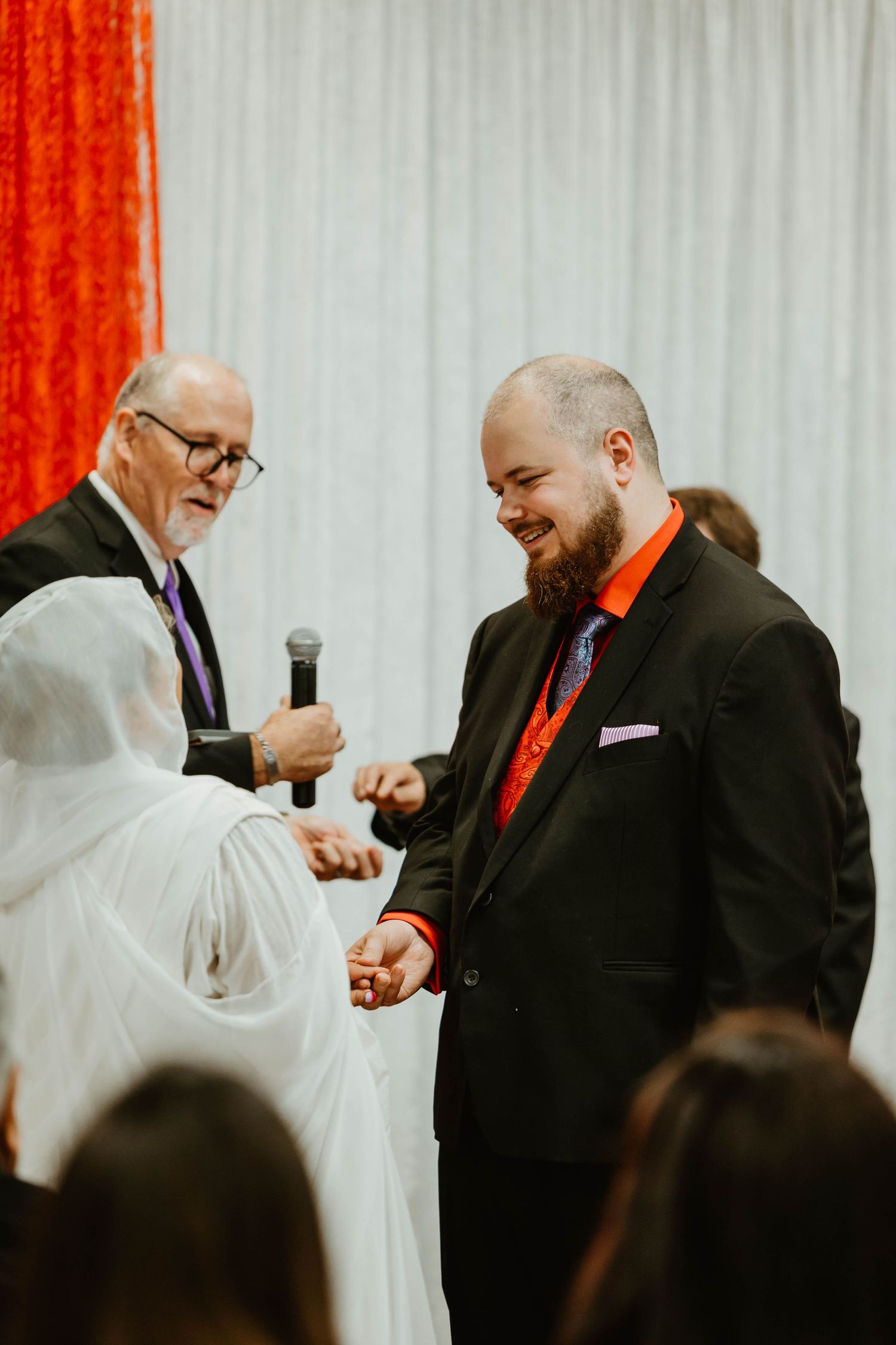 A wedding ceremony in progress with white and red decor as officiant with microphone conducts the service.