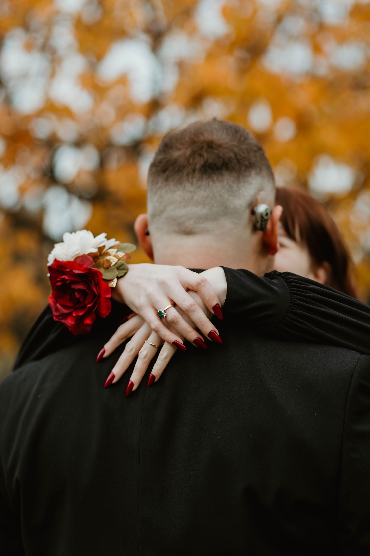 A close-up embrace showing red nails, a floral corsage and engagement ring against fall foliage in the background.