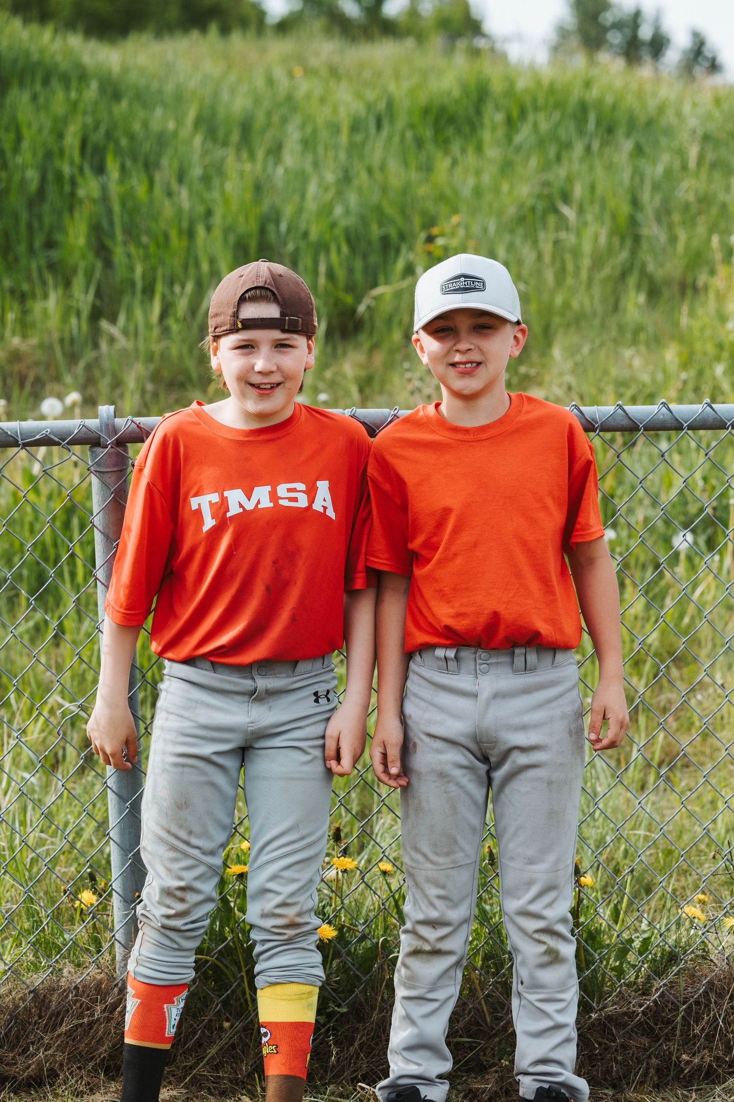 Two youth baseball players in TMSA uniforms stand together by fence.