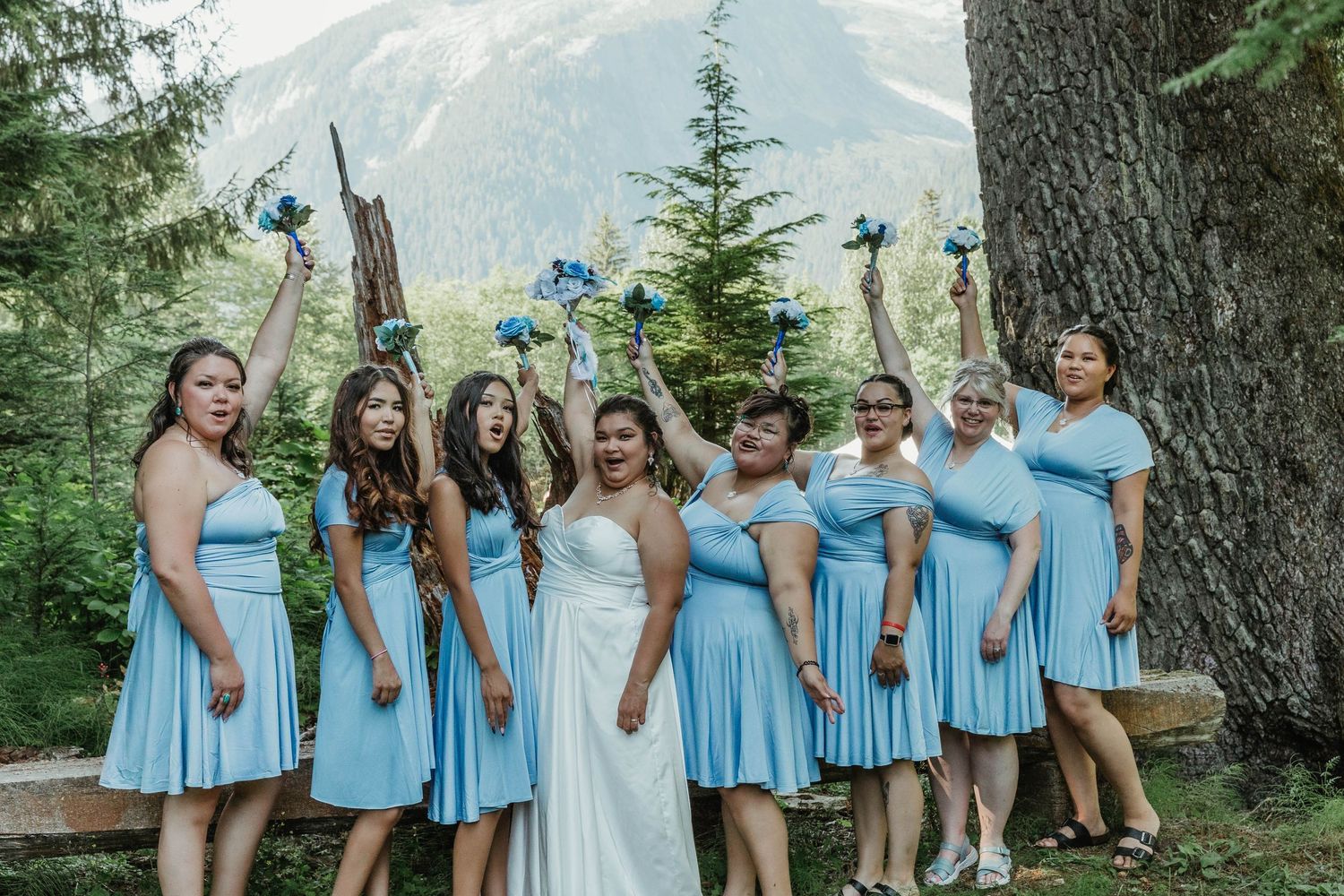 Bridesmaids in light blue dresses pose with bride against mountain backdrop holding blue bouquets.