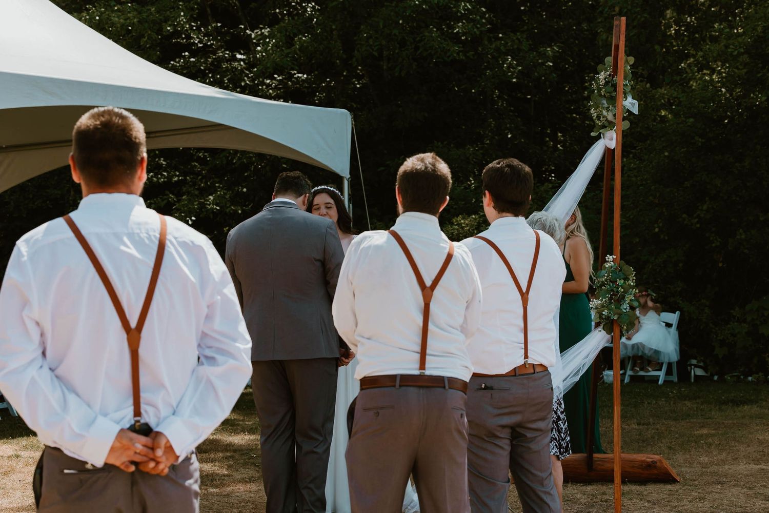 Groomsmen in suspenders watch the wedding ceremony from behind.