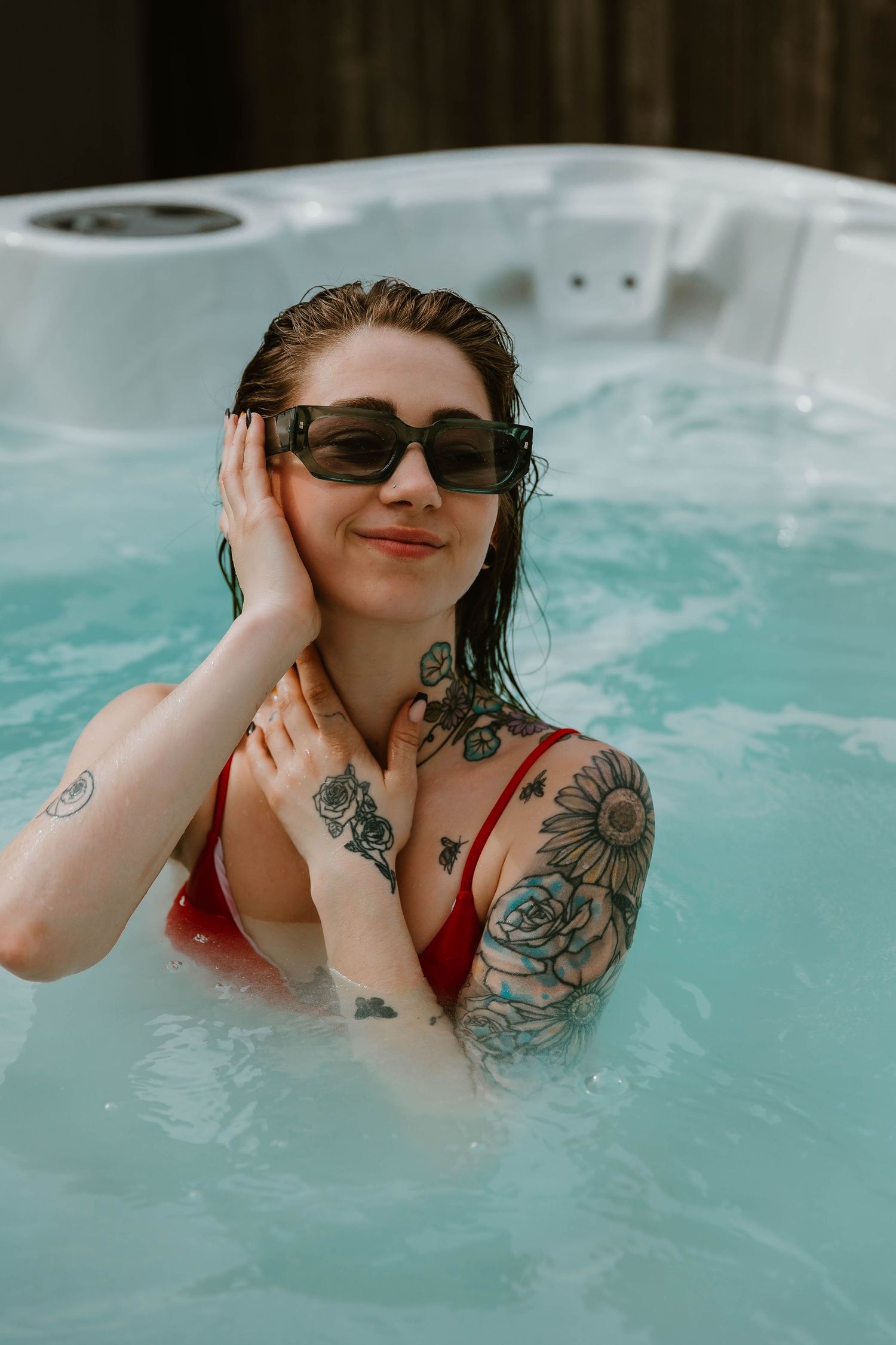 Person wearing sunglasses relaxes in blue hot tub water showing arm tattoos and red swimwear.