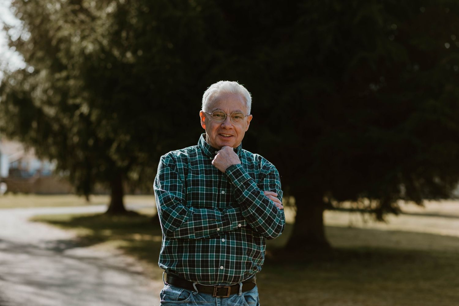 Individual wearing green plaid flannel stands outdoors in golden sunlight smiling while leaning on hand.