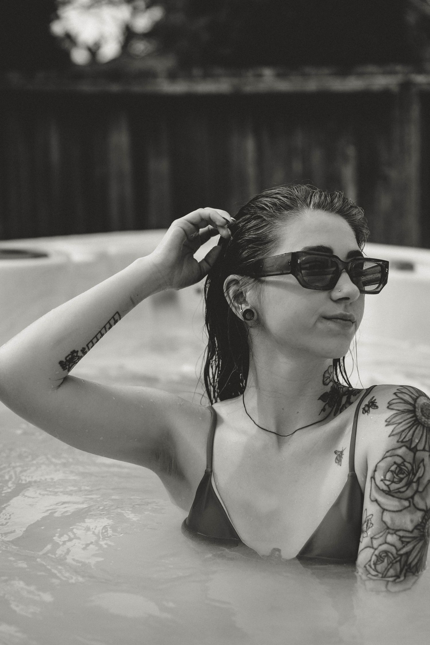 Black and white photo of someone wearing sunglasses relaxing in a spa with wet hair and visible arm tattoos.