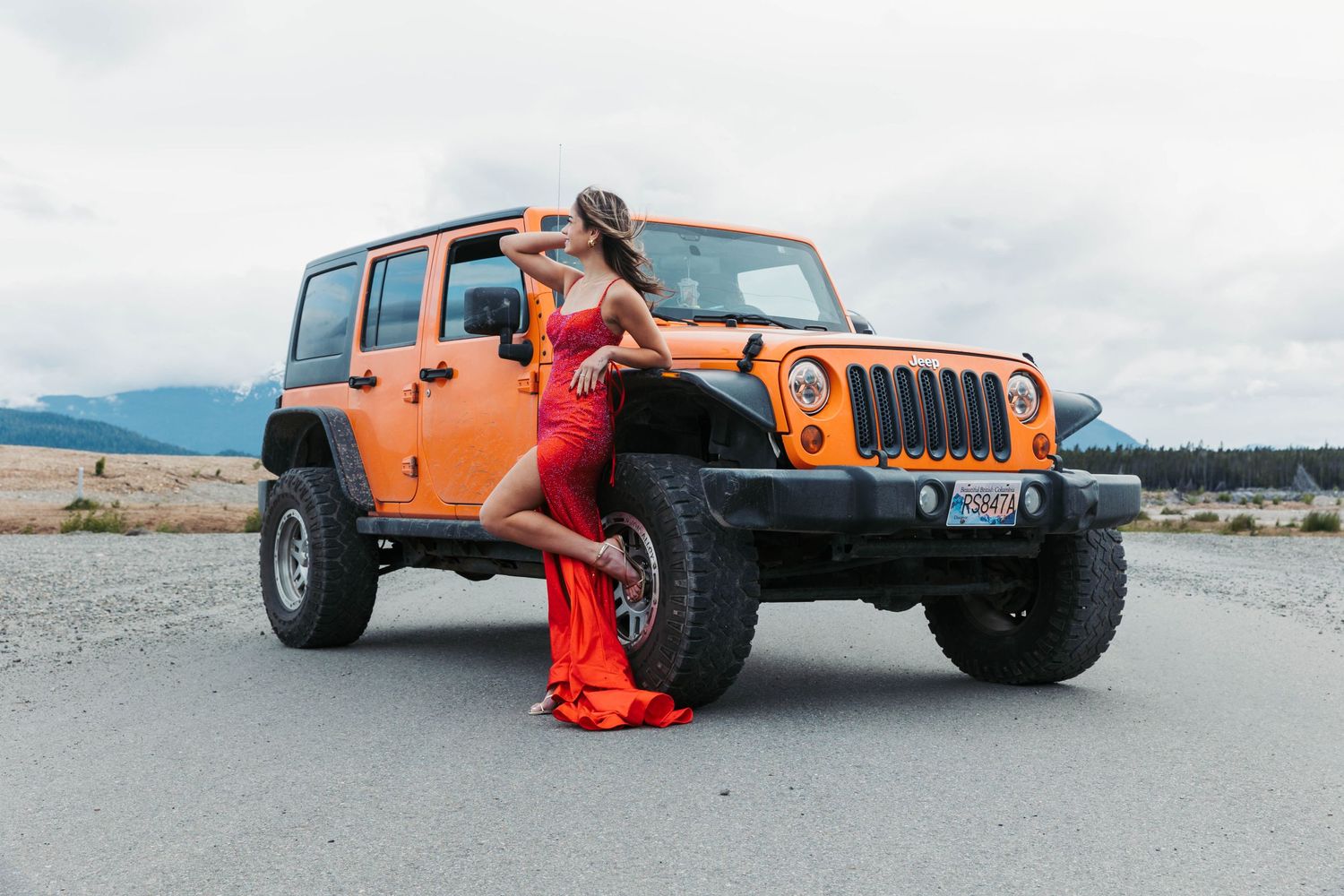 Orange Jeep Wrangler parked on a mountain road with person in red outfit posing beside it.