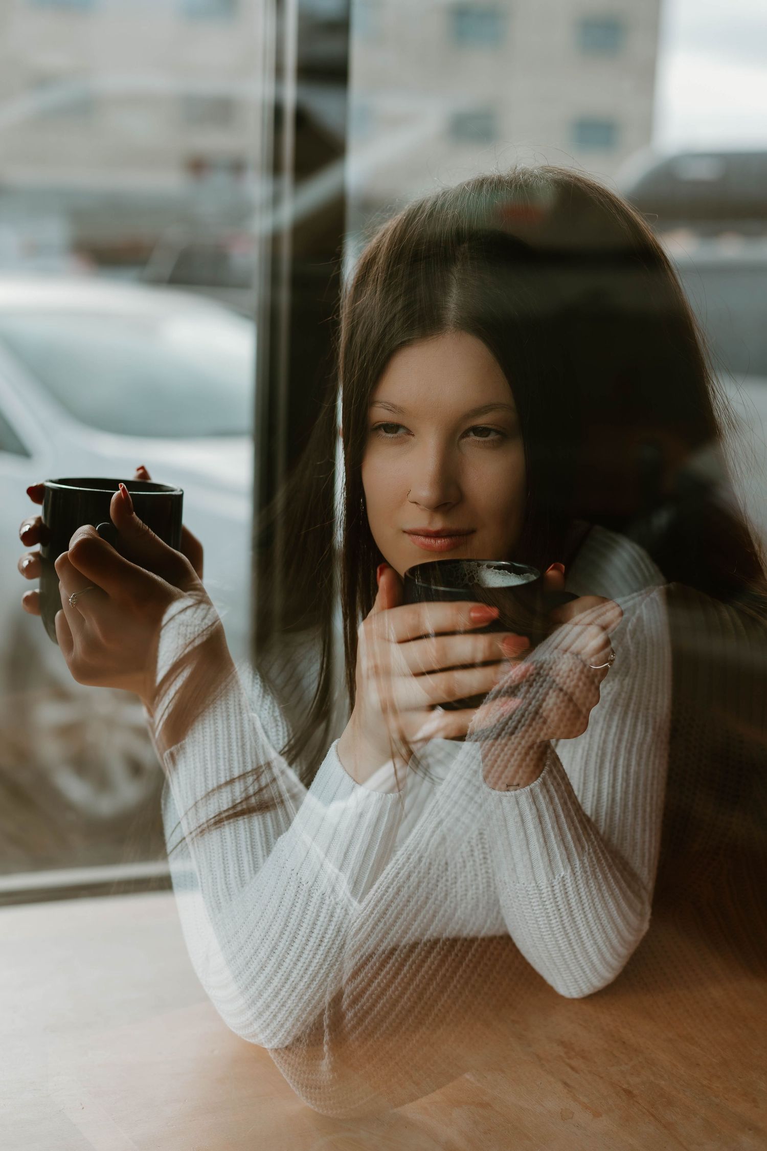 Moody coffee shop portrait through window reflection holding two cups.
