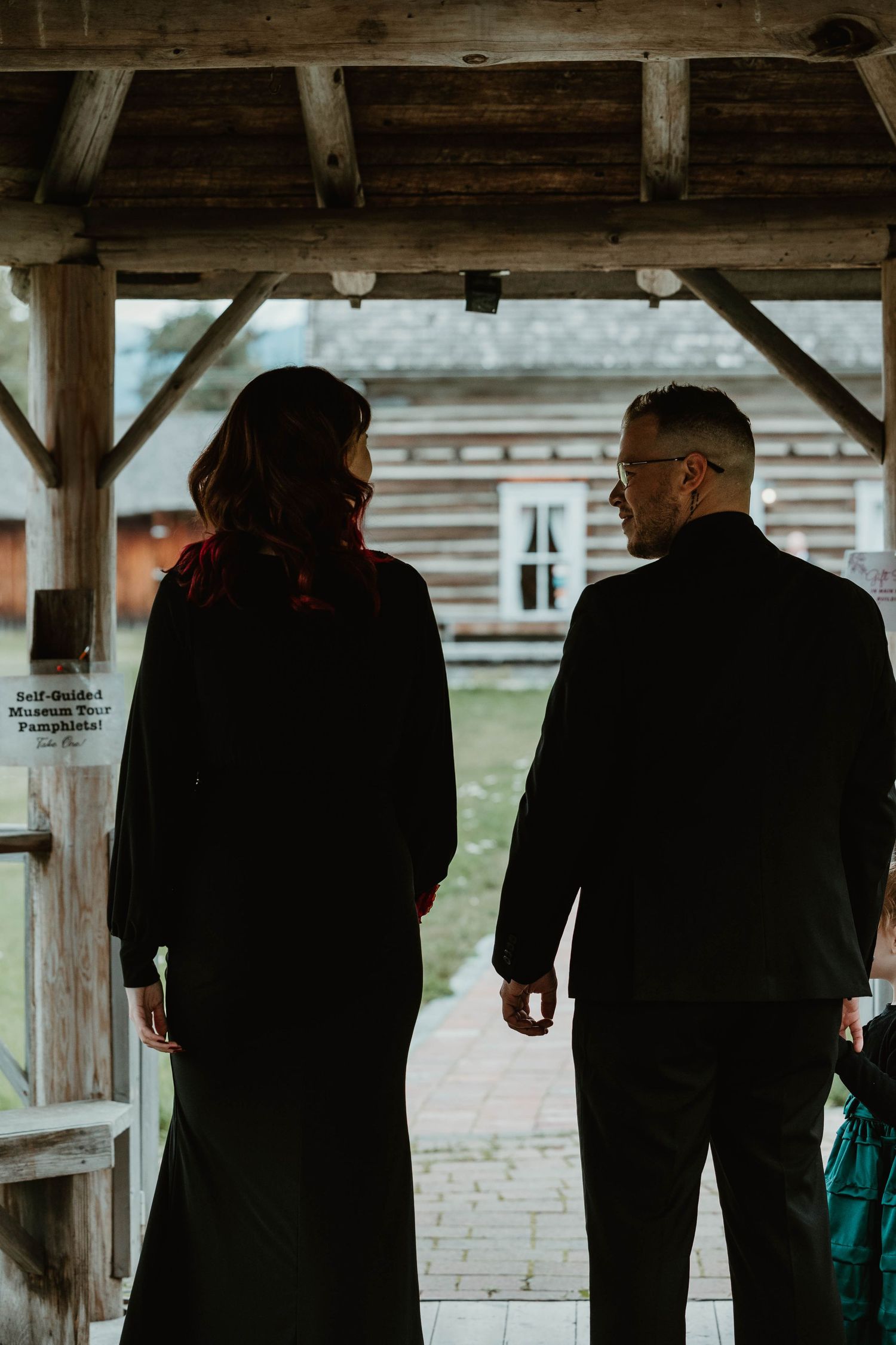 Two figures in black clothing standing under rustic wooden shelter.